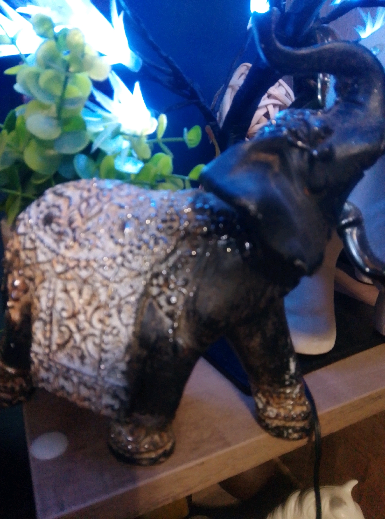 The image shows a decorative elephant figurine with intricate patterns on its body and a distressed, aged finish. The figurine is dark in color with metallic accents and appears to be made of a ceramic or resin material. It is positioned on a wooden shelf, with a blue background that may be a wall or a piece of furniture. There are also blurred green leafy plants and bright lights in the background, suggesting an indoor setting, possibly a home or a shop. The figurine's trunk is raised, giving it a dynamic pose. The overall impression is of an ornamental piece, likely part of a larger collection or display. The location in the city of Wedel, Germany is not depicted or suggested by any elements within the image itself.