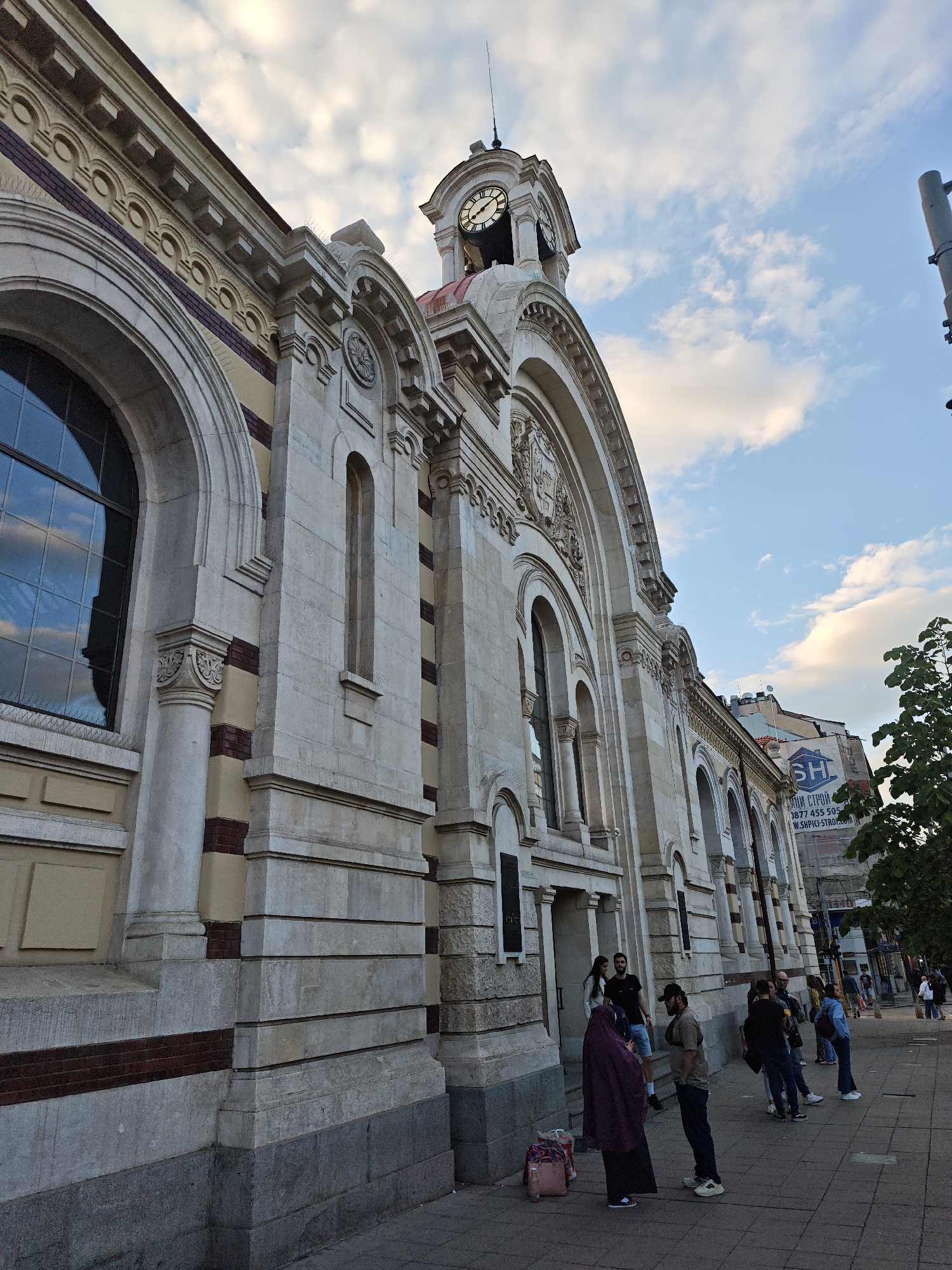 The image depicts the Central Sofia Market Hall in Sofia, Bulgaria. It is a large, ornate building with a clock tower and arched windows. The building is made of light-colored stone with some red brick accents. A group of people is standing on the sidewalk in front of the building, including a woman in a purple headscarf. The sky is blue with some white clouds. A street lamp pole is visible on the right side of the image. A building with an advertisement on the side is visible in the background on the right side of the image.