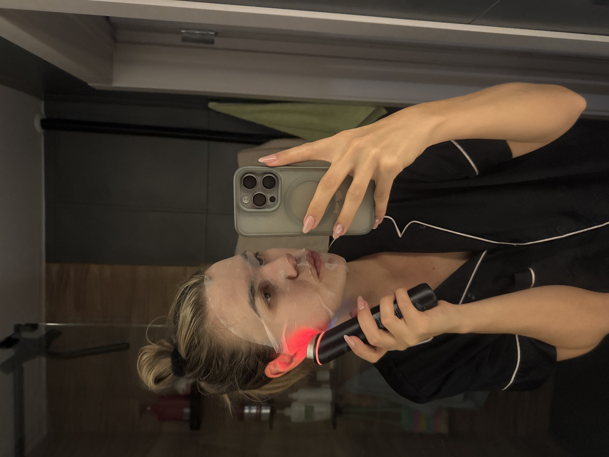 A woman is taking a selfie in a bathroom, using her phone to capture an image of herself while holding a black handheld device with a red light against her face. She is wearing a white sheet mask and a black shirt with white trim. Her hair is blonde and pulled back in a bun. The bathroom has a mirror reflecting the scene, a wooden cabinet, and possibly a towel hanging in the background. The location appears to be in a private residence rather than a public space.