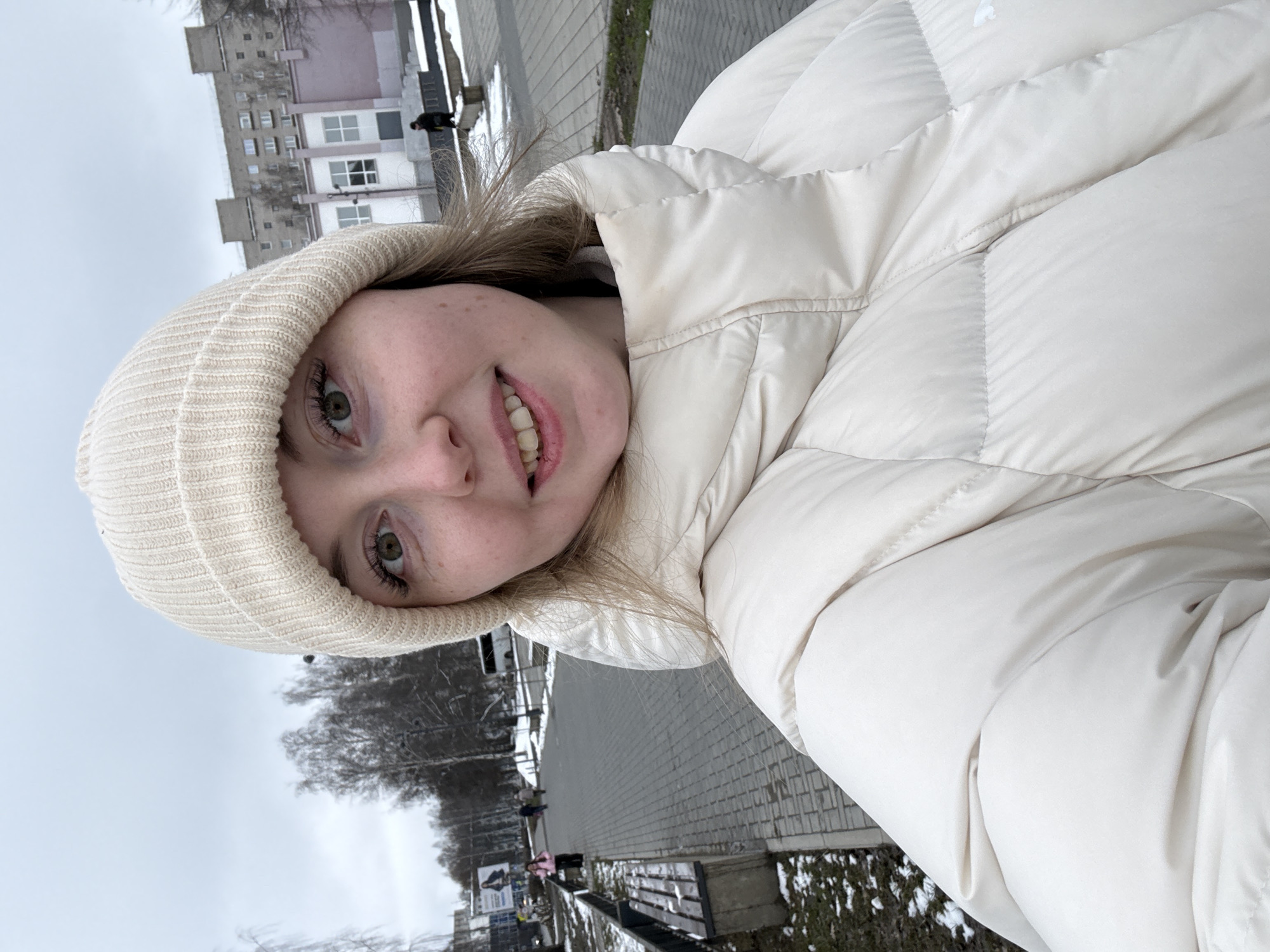 A young woman wearing a beige beanie and a puffy white jacket is shown in a close-up selfie. The background shows a city street with bare trees, sidewalks, benches, and a multi-story building with many windows. The sky is overcast.