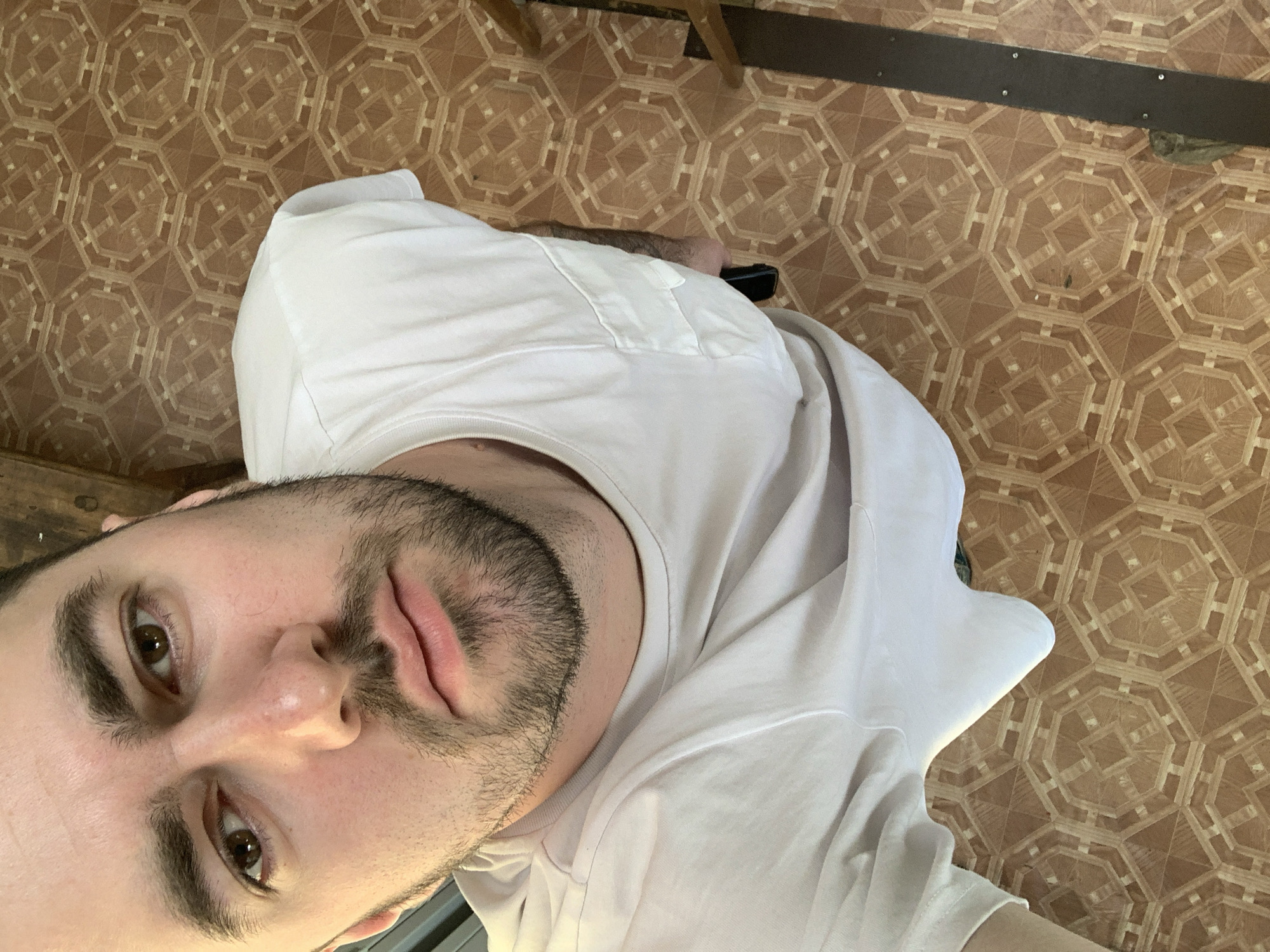 The media file is an overhead selfie of a man taken indoors in Chaikovsky, Russia. The man occupies the lower-left portion of the frame, looking directly at the camera with a neutral to slightly pensive expression, his lips slightly parted. He has dark hair, pronounced eyebrows, and a short, dark beard. He is wearing a plain white t-shirt with a visible patch pocket on the chest.
The background primarily features a brown floor covered with a distinctive geometric pattern resembling tessellated octagons and squares, possibly linoleum or patterned tile. In the upper part of the image, glimpses of dark wooden or metal furniture or structural elements are visible, including a narrow, dark beam with rivets and a broader wooden piece. The lighting suggests it is daytime. No text is visible in the image. The photo is oriented at an angle, as if taken from above and rotated counter-clockwise.