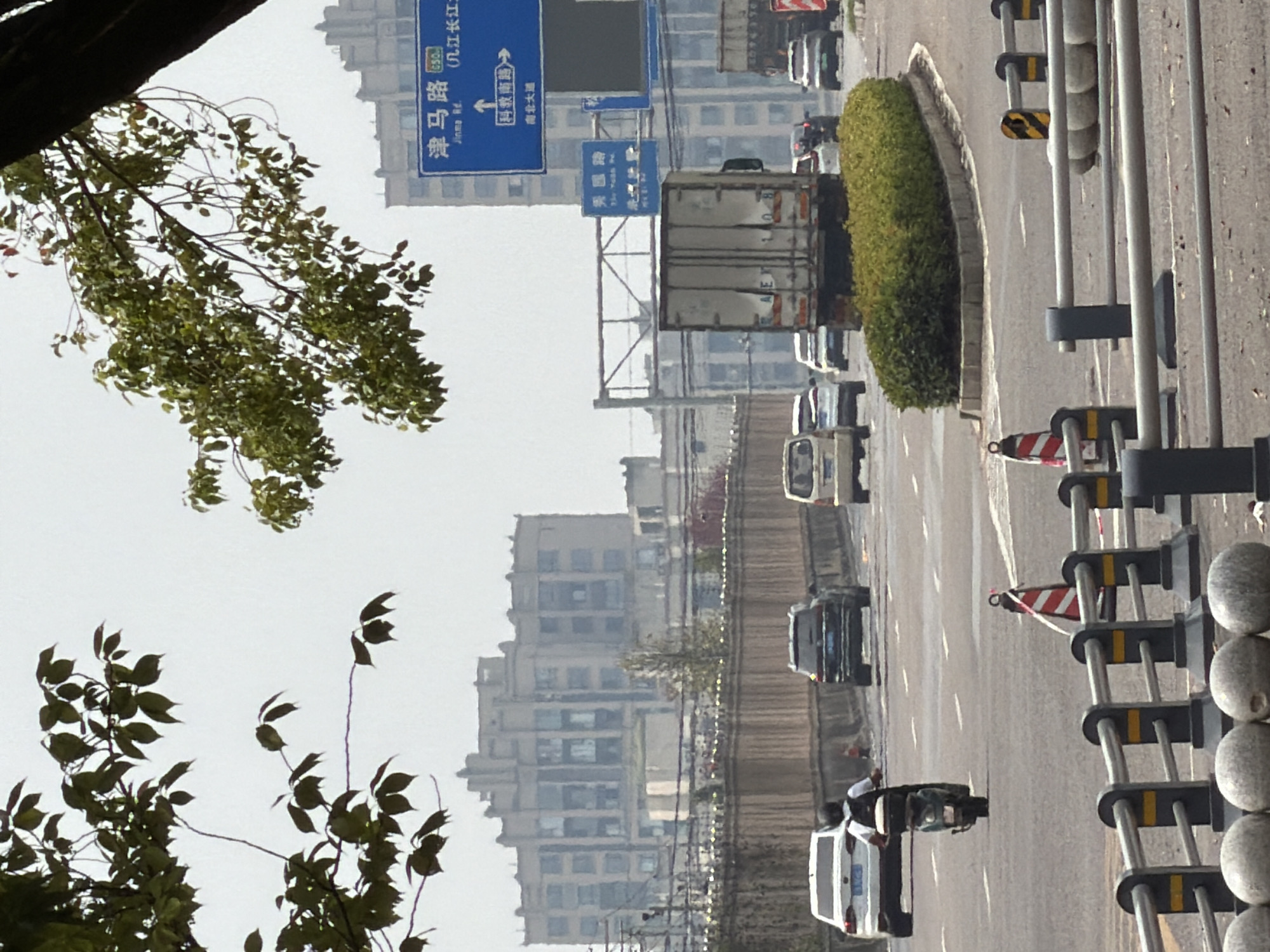 This is a photograph taken in the city of Jiangjin, China, showing a busy road intersection under a hazy sky. In the foreground, a motorcycle rider wearing a black helmet and a backpack is heading away from the camera. Behind the motorcycle, several cars are visible on the road, including a white sedan to the left and a dark SUV in the middle lane. Traffic cones and metal barriers line the road. In the middle ground, a row of neatly trimmed green bushes separates the lanes of traffic. A large white box truck is parked to the right, and a red truck is visible further in the distance. Overhead, blue and white traffic signs with Chinese characters are mounted on poles. The background features large, multi-story buildings with intricate architectural details and many windows, suggesting an urban environment. Green leaves from a tree hang into the top left portion of the frame, obscuring some of the sky and buildings. The overall scene depicts a typical urban traffic scenario with signs indicating local road names.