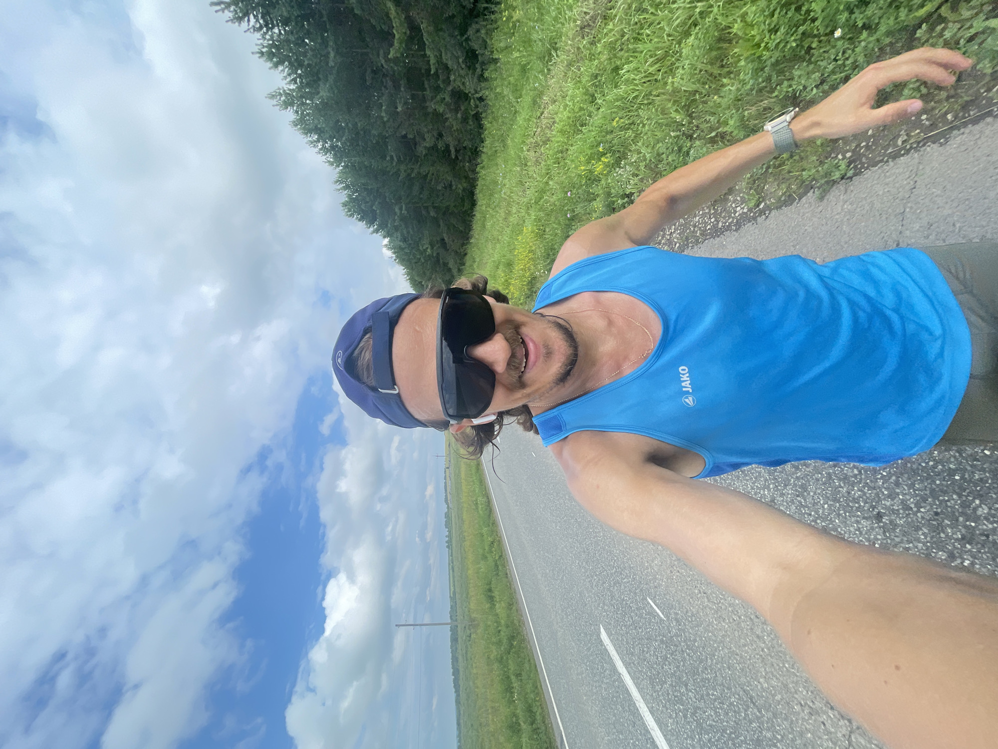 A selfie shows a man with a mustache and beard, wearing sunglasses, a blue tank top, and a blue baseball cap, running on a paved road. He has earbuds in his ears and a watch on his left wrist. The road is bordered by a grassy field and a dense forest. The sky is partly cloudy. The location is the outskirts of the city of Perevoznoe, Russia.
