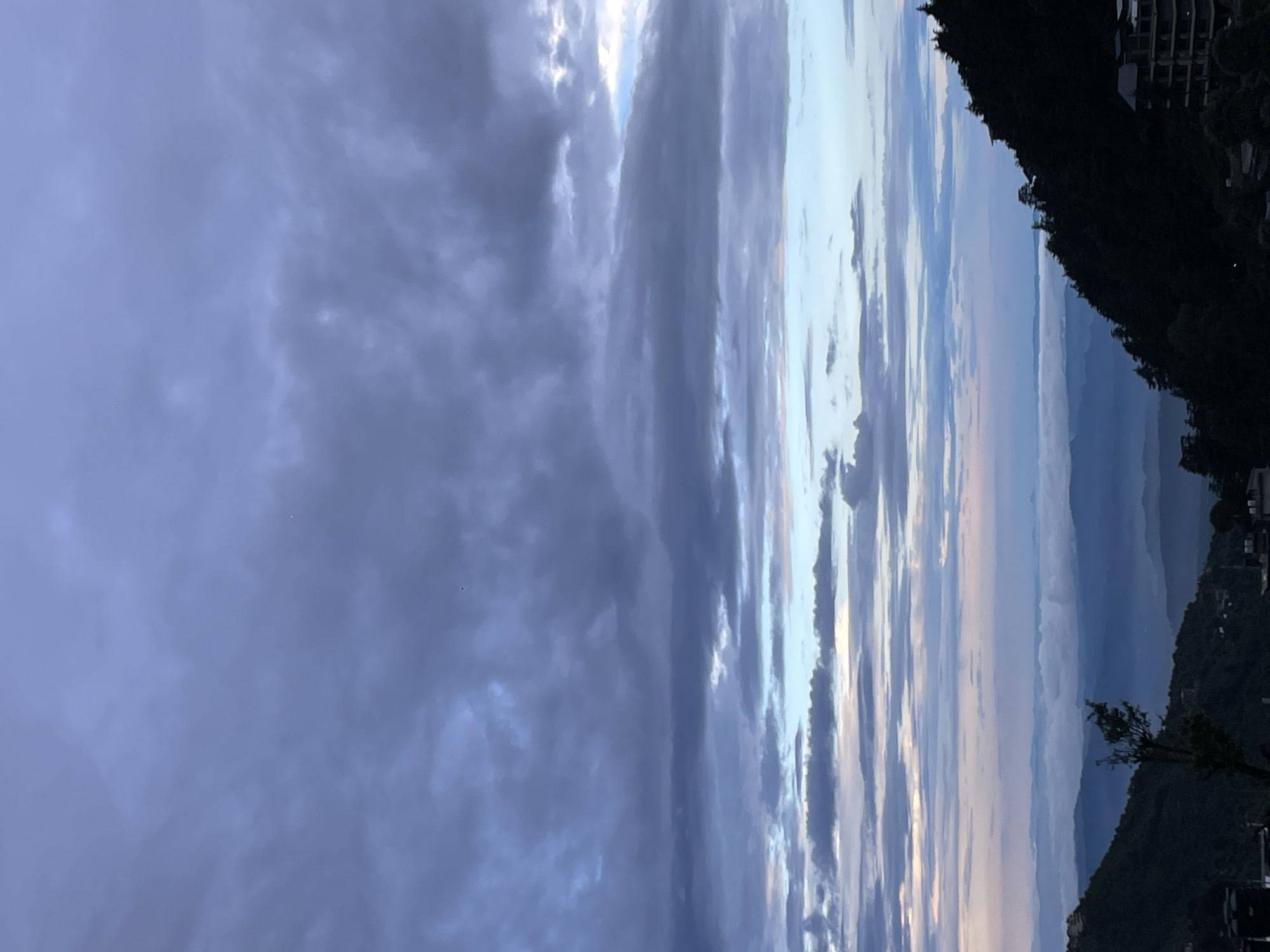 A wide-angle, vertical shot captures a panoramic view of mountains and clouds, possibly at dusk or dawn, from an elevated vantage point in Bhagsu, India. The sky dominates the upper portion of the image, filled with a vast expanse of dramatic, layered clouds in shades of gray, blue, and hints of pink. The clouds are textured and uneven, with some appearing thick and dark while others are thinner and more translucent.
Below the sky, a hazy vista of rolling mountains stretches into the distance. The mountains are rendered in soft blues and purples, with layers of mist or fog obscuring the details of further ranges. Some of these distant peaks are capped with clouds, creating a surreal, dreamlike effect.
The foreground is defined by the steep, dark slopes of a mountain covered in dense pine trees. On the right side of the foreground, a modern, multi-story building with visible windows and balconies is partially silhouetted against the sky. The building appears to be under construction or recently completed, with its dark facade contrasting sharply with the lighter tones of the sky and distant landscape. On the left side of the foreground, the dark slope continues, with a few buildings or structures visible at the base, and a solitary, slender tree stands prominently in the middle ground, its branches reaching towards the sky. The overall impression is one of a serene, expansive natural landscape juxtaposed with human habitation.