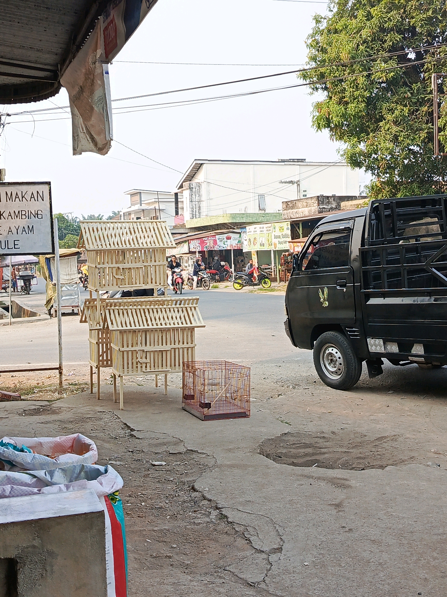 The image depicts a street scene in Megang Sakti I, Indonesia. In the foreground, there are several large sacks, likely containing goods, placed on the cracked concrete ground. To the right of the sacks, a small, ornate birdcage with birds inside is visible. Behind the birdcage, a black pickup truck is parked. In the middle ground, several wooden birdhouses, stacked and supported by legs, are displayed. A street with passing motorbikes and a few pedestrians is visible in the background. Further back, there are buildings, including shops with signs and houses, along with a large tree. Overhead wires are strung across the scene. The sky is overcast.