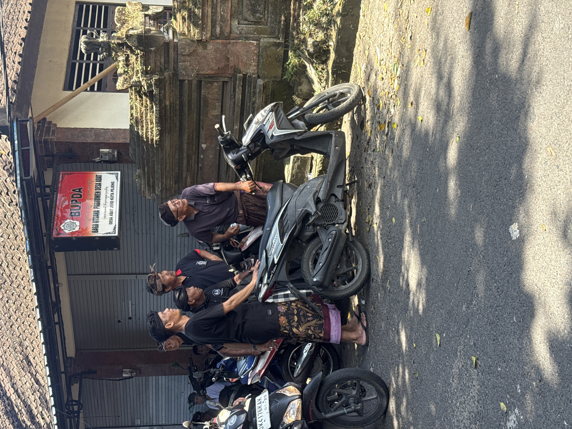 The image captures a group of men gathered on the side of a street in Pejeng, Indonesia. They are near a building identified by a sign as "BUPDA BAGA UTSAHA PRODUREN DESA ADAT," indicating a local business owned by the customary village community. The building has closed metal shutters.
Several motorcycles are parked along the road. The men, seemingly dressed in traditional Balinese attire including sarongs, stand near the motorcycles. Some appear to be looking upwards, possibly observing something above or in the distance. There are four visible men and multiple additional individuals are seen in the background with their motorcycles. The location is likely a street in Pejeng.
