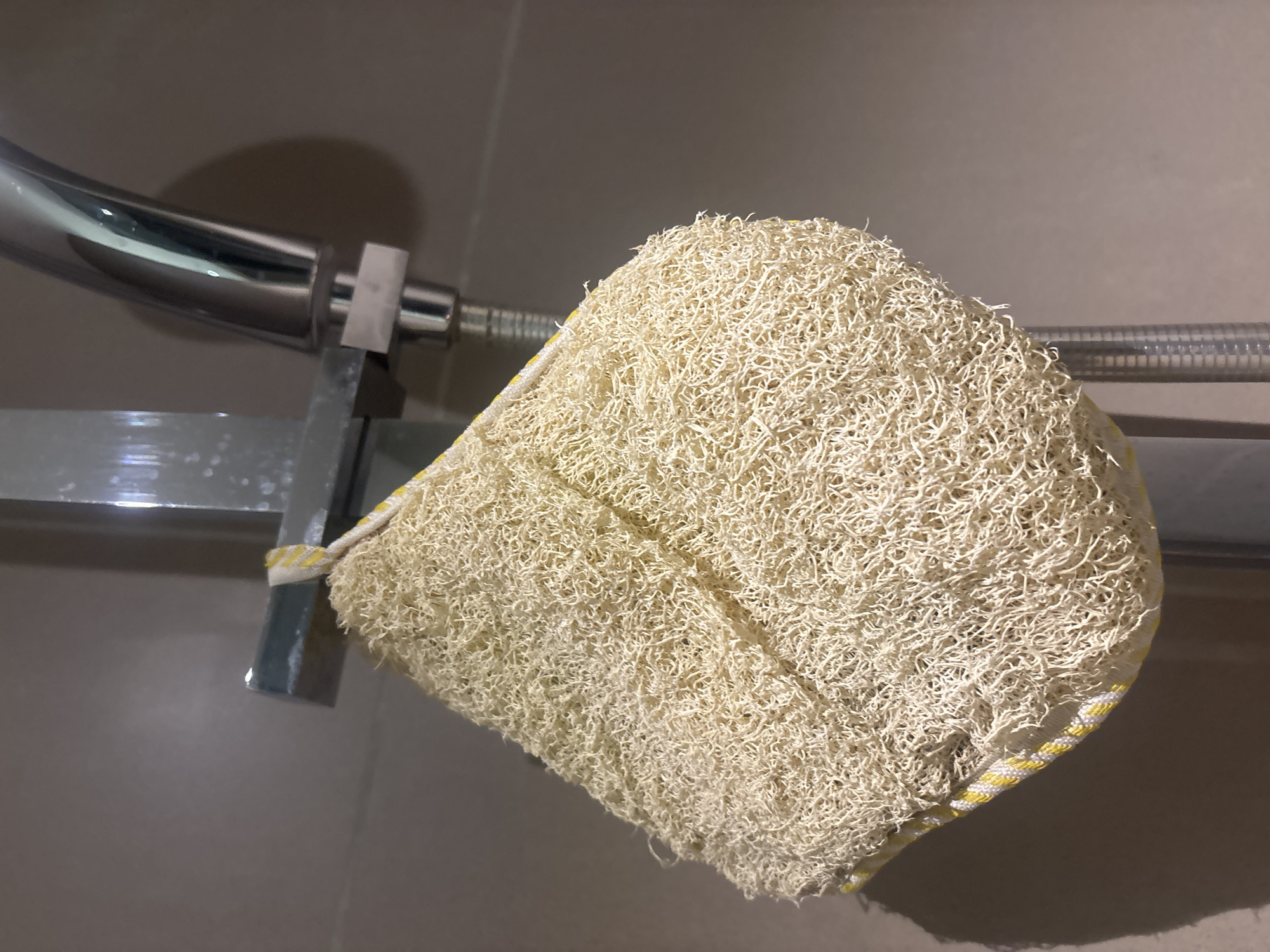 A close-up, eye-level shot shows a loofah sponge hanging from a silver shower head pipe in a bathroom. The loofah is light beige, with a woven texture and a yellow and white striped trim. The shower head is made of chrome and is partially visible. The background is a tiled wall in a neutral, greyish-brown color. There are no people or animals in the image. The lighting suggests an indoor setting, likely a modern bathroom. The image does not contain any specific landmarks or elements that would allow for an accurate determination of its location within Dubai, United Arab Emirates.