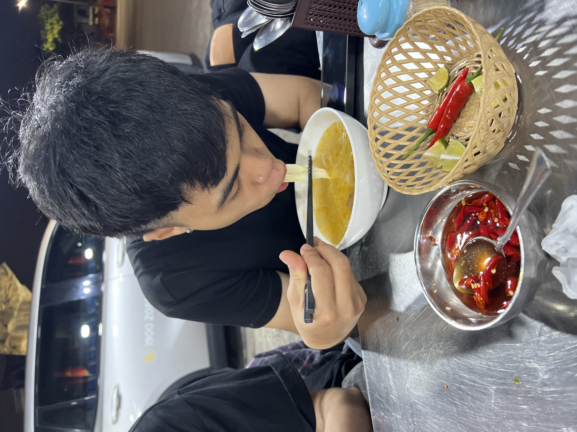 A person with black hair wearing a black t-shirt is eating noodles with chopsticks from a white bowl on a metal table. Next to the bowl is a wicker basket containing red chili peppers and lime wedges, and a metal bowl with red chili peppers in oil. The person is positioned at an outdoor restaurant table in Đà Nẵng, Vietnam; partially visible in the background is a car with the phone number "1900 2017".