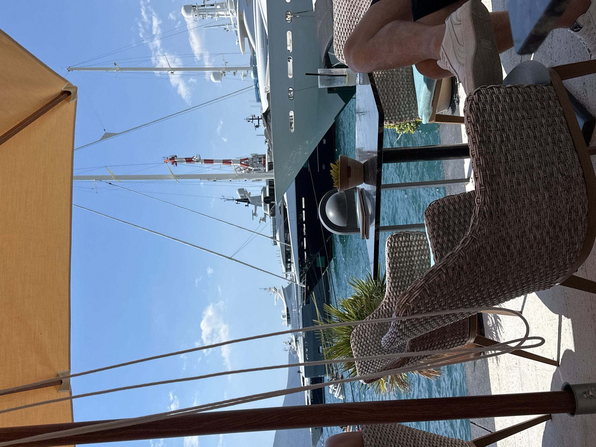 The image depicts an outdoor seating area at a cafe by the marina in Donja Lastva, Montenegro. In the foreground, wicker chairs surround a dark table. A glass of water with ice and a lemon slice, a small potted plant, and a notebook are visible on the table. A person's legs and feet, wearing white sneakers, are prominent in the lower right portion of the frame, indicating they are seated at the table. In the background, several large yachts are docked in the bright blue water under a clear sky. The marina is framed by a large, tan umbrella providing shade over the seating area. The overall scene suggests a relaxed atmosphere in a scenic coastal location.