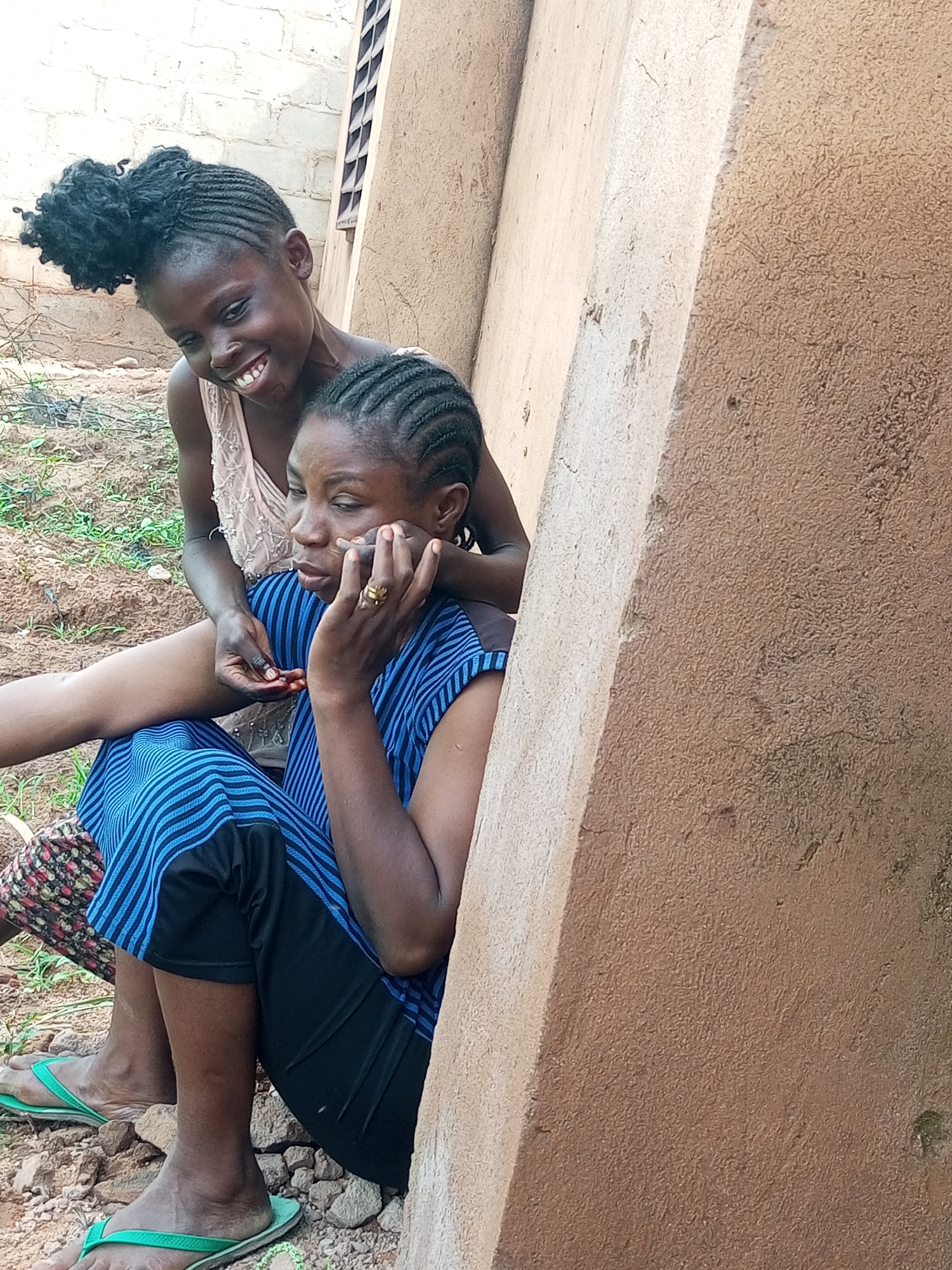 Two young Black females are seated outdoors, leaning against a textured, light brown wall. The female in the foreground, on the right, is wearing a blue and black striped dress and black pants. She has her left hand on her cheek and is wearing a gold ring on her right hand. Her hair is braided. The female behind her, on the left, is wearing a light-colored sleeveless top and is leaning over, smiling and looking down. Her hair is in a bun. The ground around them is rough, with scattered rocks and sparse vegetation. In the background, a light-colored brick wall is visible. The scene appears to be in a rural or semi-rural area of Tempure, Nigeria.