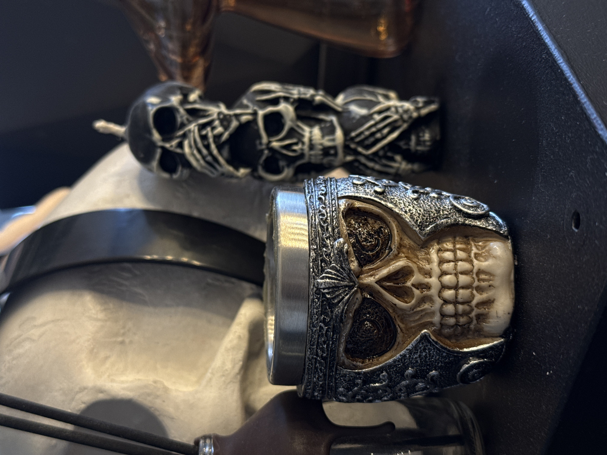 The image shows two skull-themed objects. In the foreground, there is a skull ring, with a metal band. Behind it and slightly above is a row of skulls arranged in a line. The image was likely taken indoors, based on the lighting and surroundings. It is not possible to discern the location in Krasnoyarsk, Russia from this image.