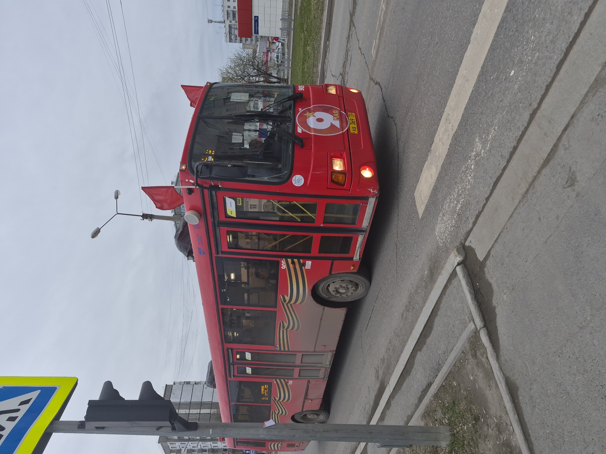 The media file captures a red public bus on a paved street in Tobolsk, Russia, under an overcast sky. The bus is heavily decorated, suggesting a commemoration of Victory Day, observed annually on May 9th.
The prominent subject is the long red bus, which features two red flags mounted on its front roof corners. A large circular emblem on the front displays a stylized number "9" and the Cyrillic word "МАЯ" (MAY), directly referencing May 9th. The bus also shows a route number "905" on its windshield and a Russian license plate "У228АВ 369 72". Its headlights and indicator lights are illuminated.
Along the side of the bus, a significant graphic resembling the black and orange striped Ribbon of Saint George is visible, a widely recognized symbol of military valor in Russia, particularly associated with Victory Day. The bus appears somewhat dirty or dusty, especially near its wheels. Passengers can be seen through the windows, indicating it is an active public transport vehicle.
The scene includes typical urban infrastructure: an asphalt road with white markings for a crosswalk, concrete curbs, a grassy verge, and power lines overhead. In the foreground, a blue and white pedestrian crossing sign and a traffic light are visible. Bare trees and multi-story buildings populate the background. The overcast weather and bare trees suggest the photograph was taken in late winter or early spring, consistent with preparations or celebration around early May.
