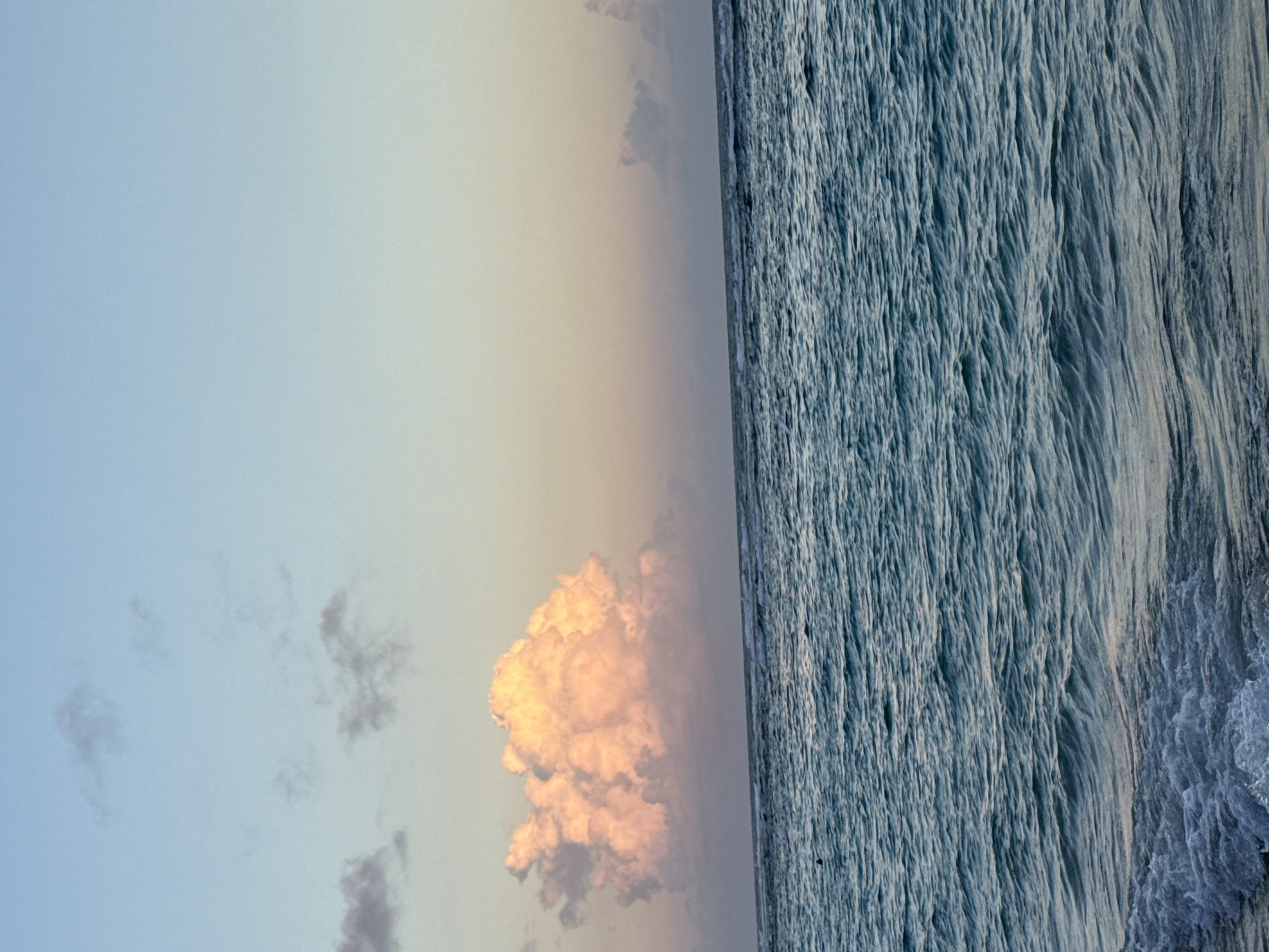 A landscape photograph shows an ocean scene with a sky containing a single large, fluffy, pink-hued cumulus cloud. The cloud is situated in the lower middle of the frame, illuminated by the setting sun. Smaller, indistinct cloud formations are visible to the left. The sky transitions from light blue to a soft golden hue, likely due to the sunset. To the right, the ocean’s surface is textured with numerous small waves or ripples, appearing light blue. The horizon line is faintly discernible between the sky and the ocean. The composition is oriented vertically. The location is unknown.