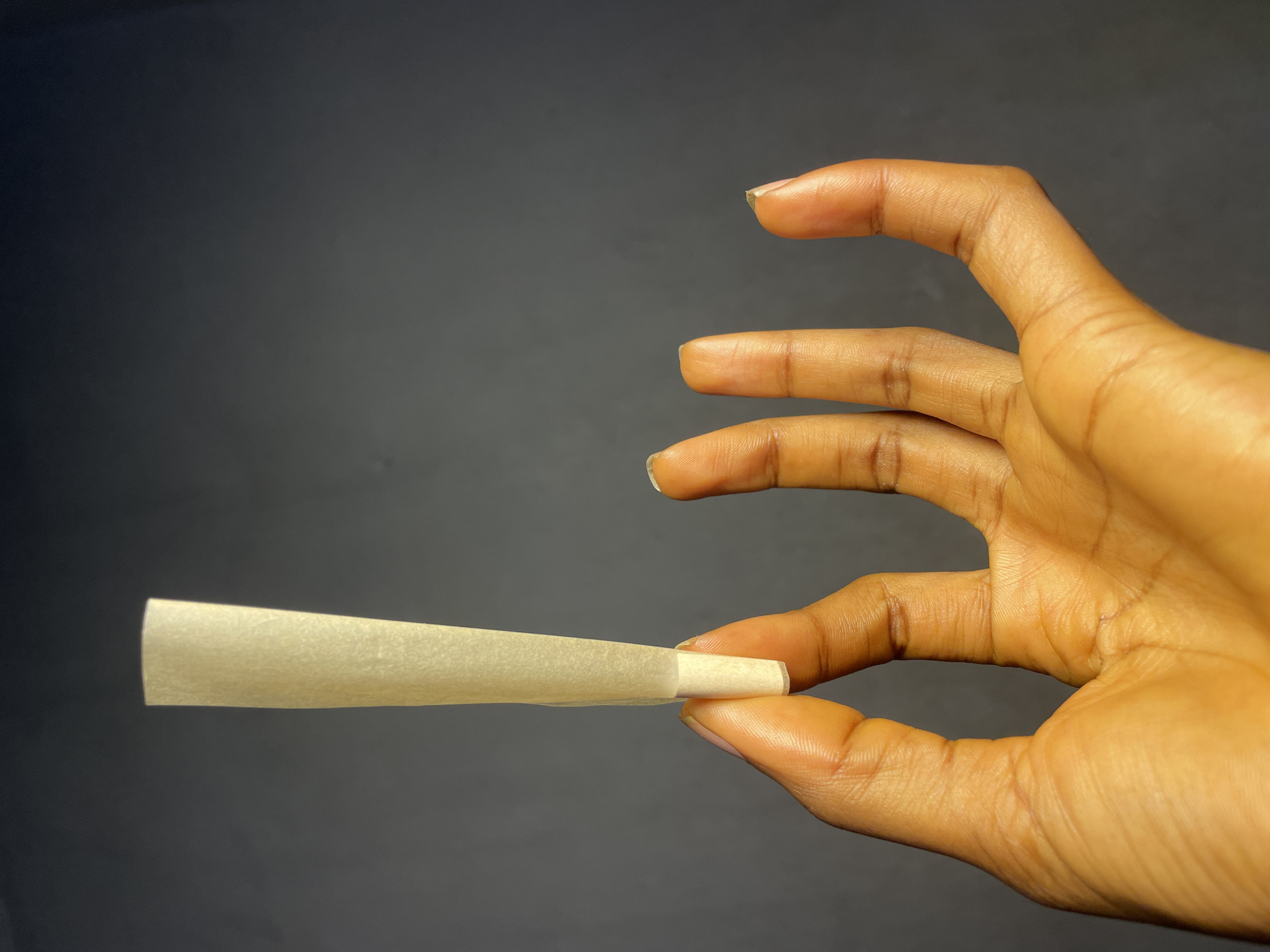 A hand holds a rolled cigarette against a dark gray background. The hand is dark-skinned and angled with the fingers slightly curved. The rolled cigarette is held between the thumb and forefinger, pointing towards the left. The location, Nkpor, Nigeria, is not discernible from the visual content.