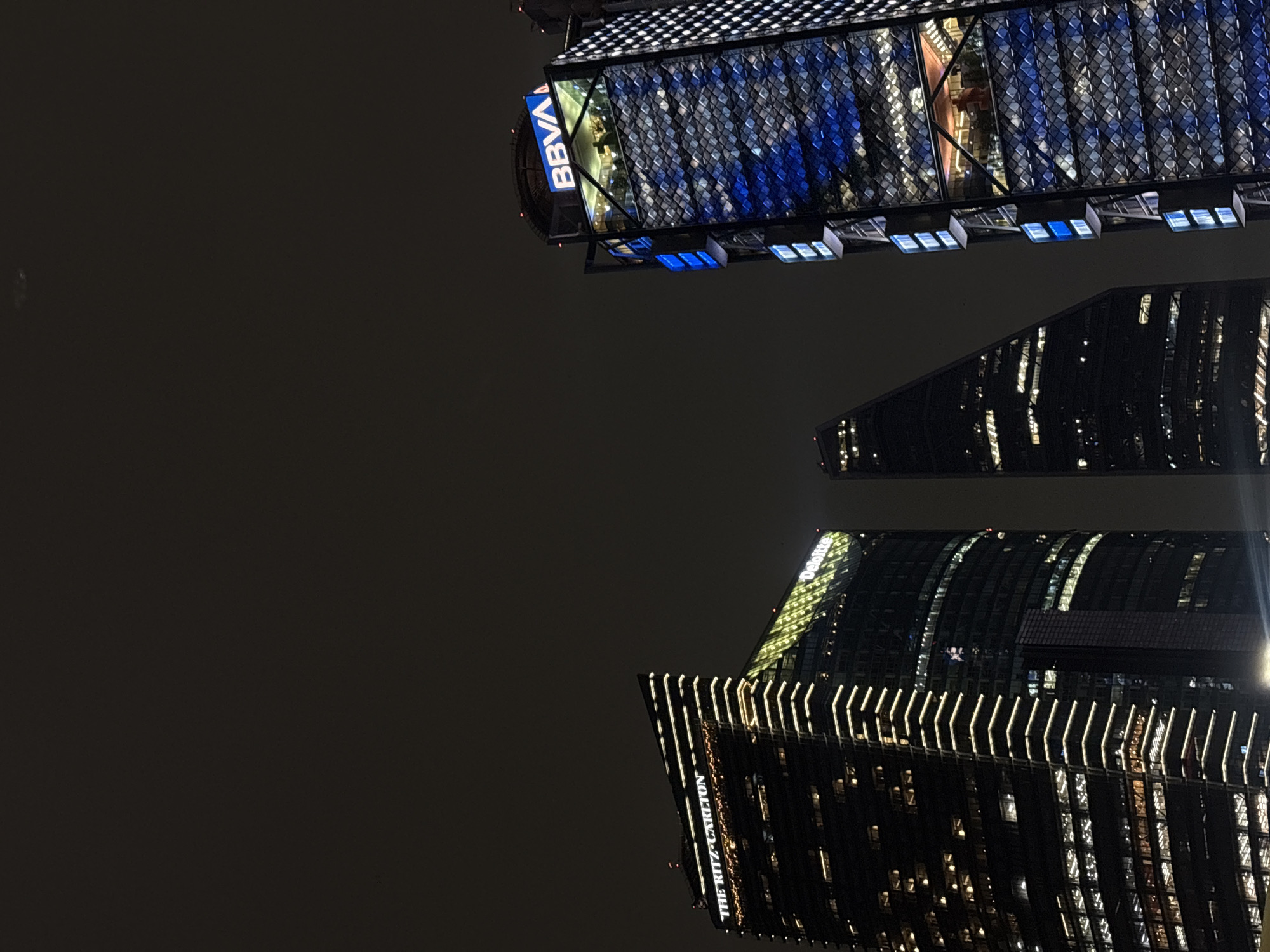An upward-angled nighttime photograph captures three illuminated skyscrapers against a dark, overcast sky. The prominent building on the left features a brightly lit "THE RITZ-CARLTON" sign at its apex and "Deloitte" illuminated in yellow-green below it, with its facade outlined by horizontal and vertical white-yellow LED strips. Directly to its right, a dark, triangular-shaped skyscraper is partially visible, showing scattered lit windows. On the far right, a distinct skyscraper displays a blue "BBVA" logo at its summit. Its facade is patterned with numerous tessellated panels, primarily illuminated with blue and white lights. Below the "BBVA" logo, an open-concept or glass-enclosed upper level reveals several silhouetted human figures, indicating activity within. Vertical blue light panels are visible along its structure. The overall scene depicts modern architecture in a dense urban environment at night. The identifiable buildings place the location in Mexico City, Mexico.