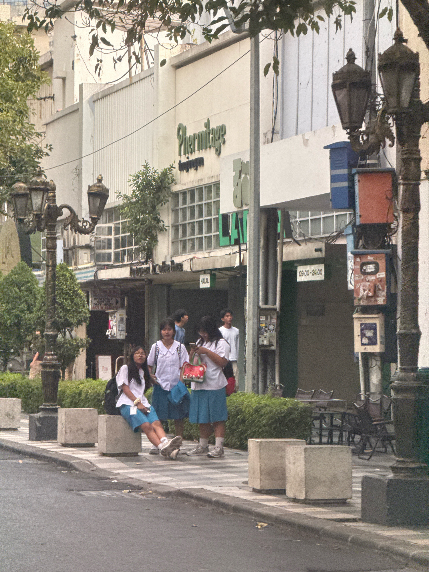 In Surabaya, Indonesia, on a street, three young women, likely students based on their uniforms, stand on a sidewalk. One woman in the foreground, wearing glasses and a white shirt and blue skirt, sits on a concrete planter. To her right, another woman in a similar uniform stands, holding a red handbag with a floral pattern. A third woman in uniform stands further to the right. Behind them, a building with the signage "Hermitage" is visible. Several lampposts, some ornate, line the street. Tables and chairs are set up outside a cafe or restaurant. The scene appears to be set in the late afternoon or early evening.