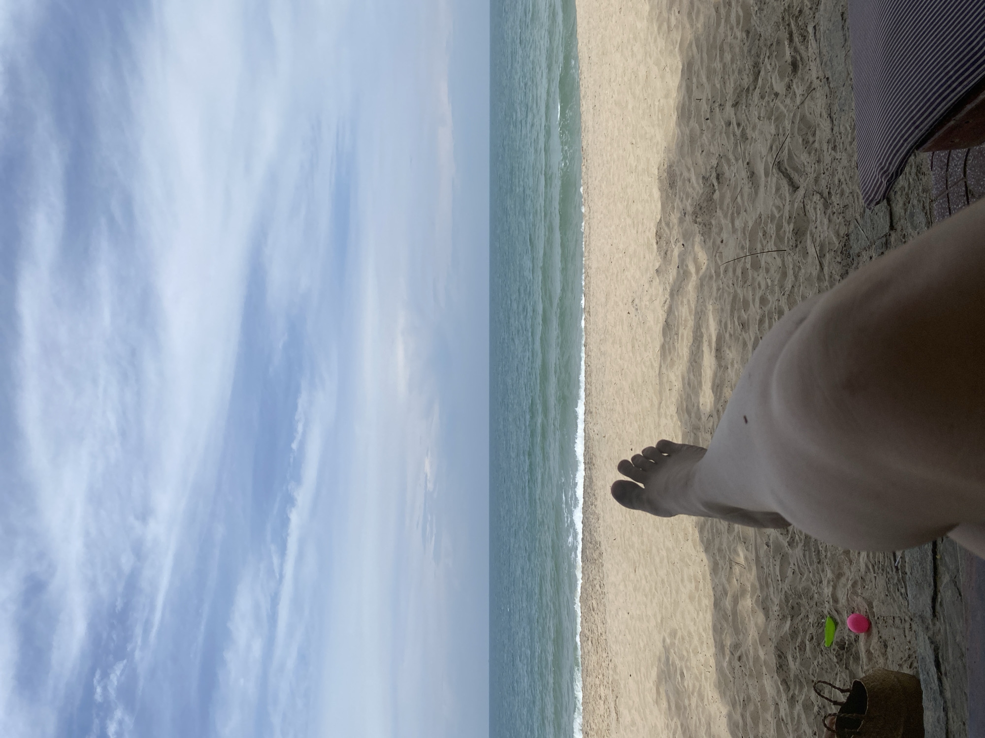 The image shows a first-person perspective of a person relaxing on a beach. The person's legs and feet are in the foreground, with their right leg bent and extended towards the ocean. The beach sand is light brown and appears dry, with some scattered shadows. Beyond the sand is the ocean, with gentle waves breaking on the shore. The water is a clear turquoise color. In the distance, the horizon line separates the ocean from a hazy sky with scattered clouds. A straw bag and some small toys, a pink ball and a green shovel, are visible on the sand to the left. A striped cushion is partially visible in the lower right corner. The scene suggests a peaceful and relaxing day at the beach. While the image does not provide specific landmarks to identify Phu Thuy Ward, Vietnam, the setting is consistent with a coastal location in the region.
