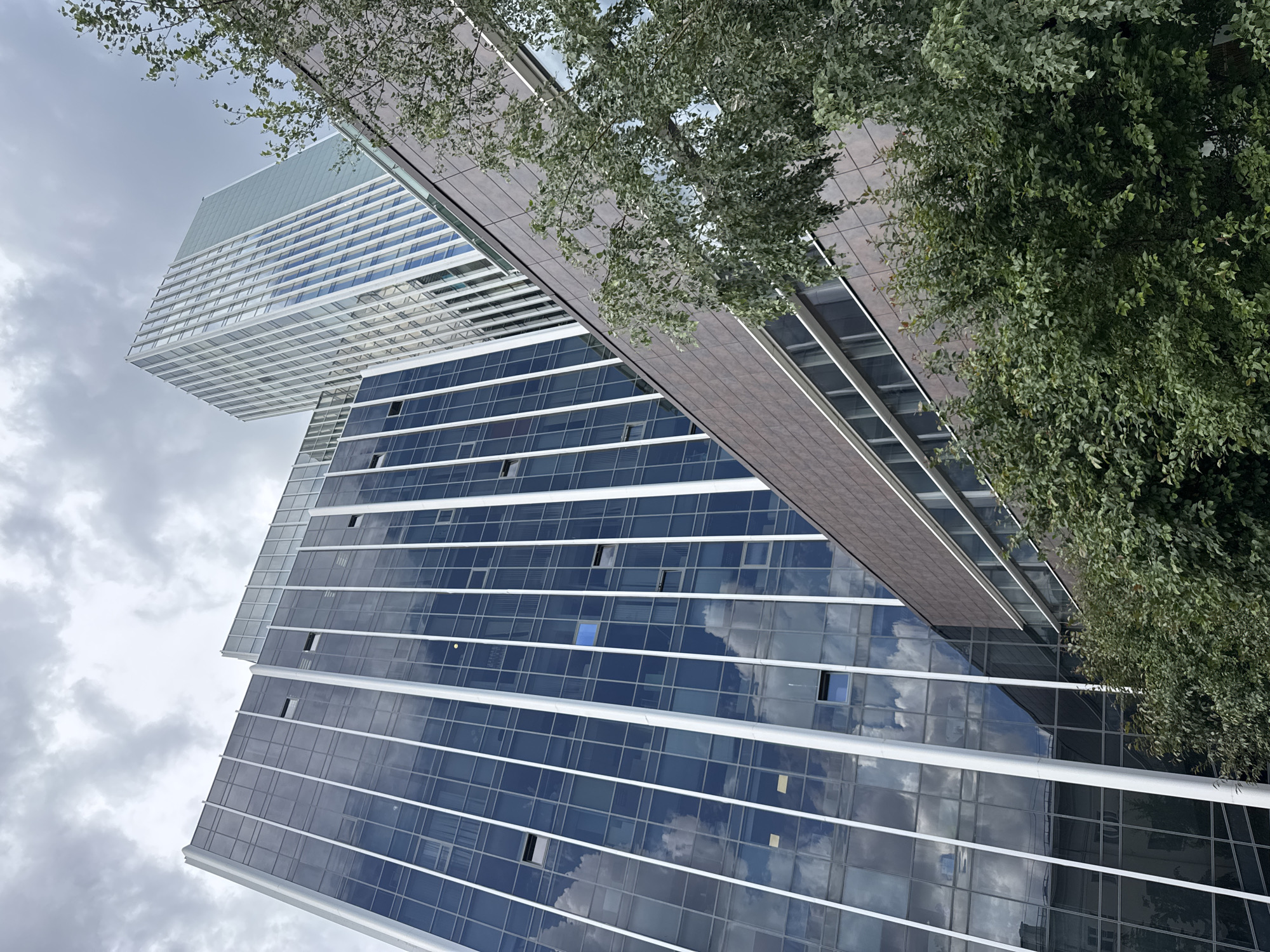 The image shows a modern architectural scene with two buildings and foliage. One building is tall and slender with a facade of blue-tinted glass windows and white horizontal accents. The other building, partially visible, has a lighter facade with horizontal white lines and a greenish top section. A connecting structure with a dark tiled surface spans between the buildings, with lush green leaves of a tree obscuring parts of it. The sky is overcast with clouds, suggesting a daytime setting. The reflection of the sky and clouds is visible in the glass windows of the lower building. The perspective is from below, looking upwards, creating a dynamic and somewhat disorienting view of the structures. No people or specific activities are depicted. The location cues are limited to the architectural style and the context provided about Yekaterinburg, Russia. The overall mood is neutral, focusing on the urban landscape and its elements.