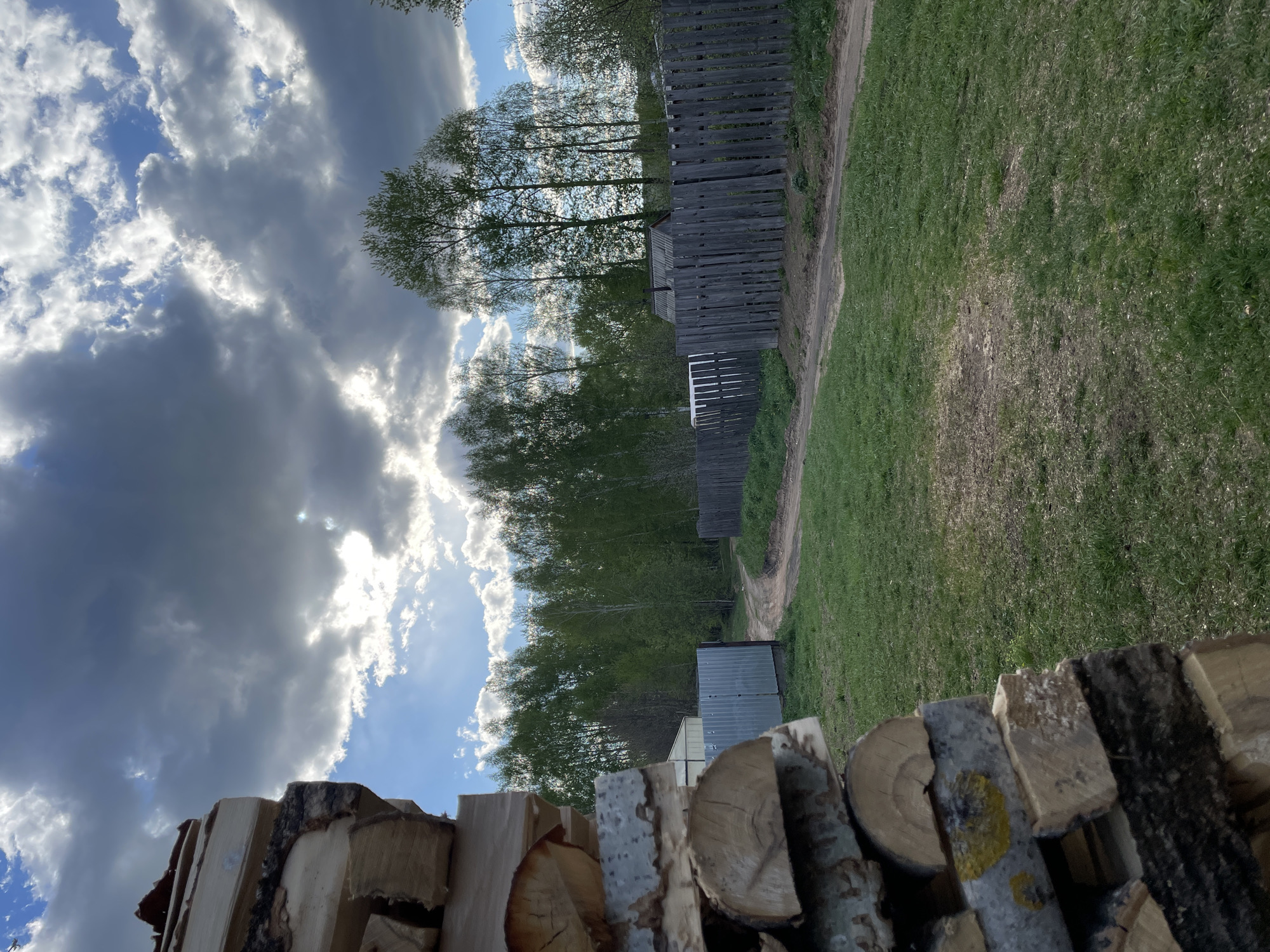 The image shows a rural scene in or near Балдейка, Russia. In the foreground is a stack of firewood, cut logs stacked neatly. Beyond the firewood is a grassy field bordered by a wooden fence. A dirt path runs alongside the fence. In the middle ground, there is a small shed-like structure. Trees are visible behind the fence and shed. The sky is dramatic with a mix of dark, heavy clouds and patches of blue sky, with the sun occasionally shining through. There are no people visible in the image.