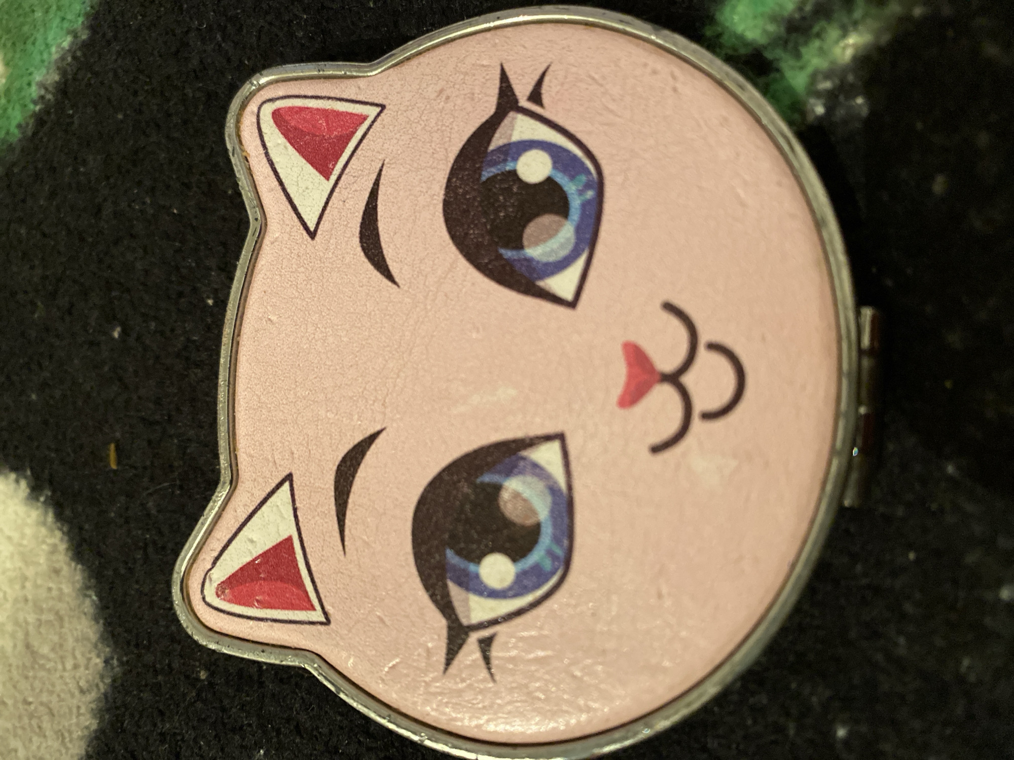 The image shows a close-up, overhead view of a pink, cat-shaped compact mirror with a silver border. The cat face features large blue eyes with black outlines and eyelashes, small arched eyebrows, and a small pink nose. Below the nose, a simple mouth is drawn with two curved lines. The inside of the cat's ears are depicted as red triangles. The compact is resting on a dark, textured surface, possibly a carpet or rug, with hints of green and white pattern visible in the background. There are no people or animals present in the image, nor is there any indication of the city of Maloarslangulovo, Russia, or any specific location. The lighting suggests it is indoors.
