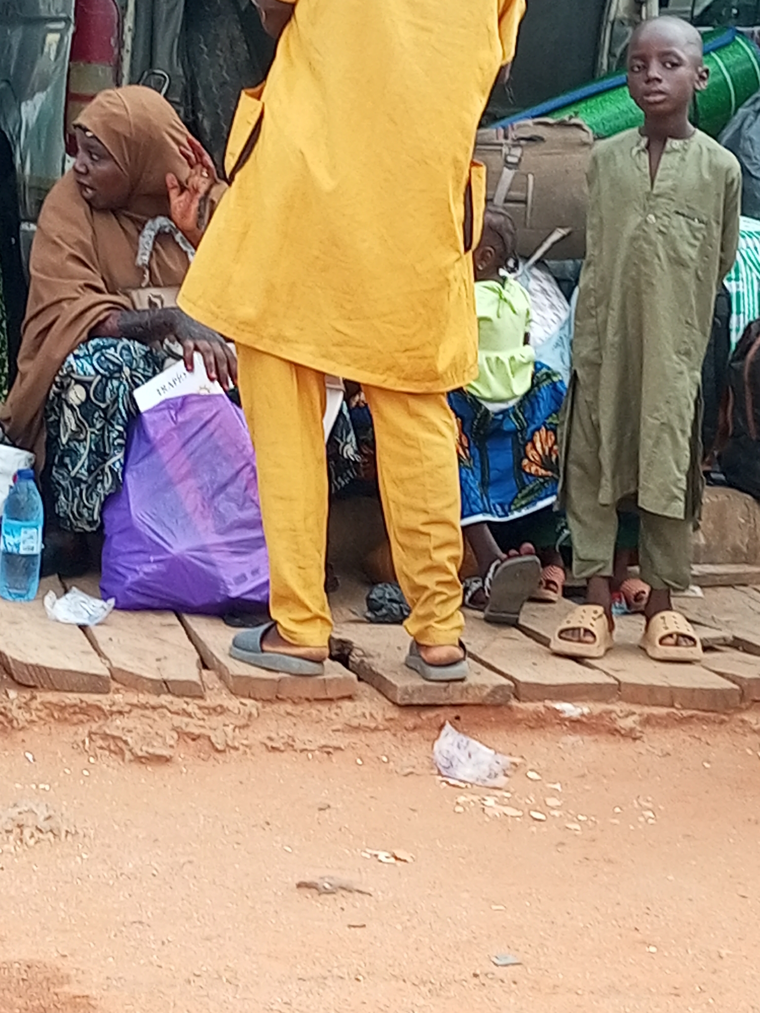 In Gombe, Nigeria, a group of people are gathered outdoors, possibly at a bus station or market, with luggage and bags. A woman in a brown hijab and patterned skirt sits on a wooden platform, holding a purple shopping bag. Next to her, a person in a bright yellow tunic and pants stands, facing away from the camera. To the right, a young boy in a green tunic and pants stands looking at the camera, while another child in a light green outfit is visible behind him. The ground is made of reddish-brown dirt, with scattered pieces of trash. The scene suggests a moment of waiting or transit.