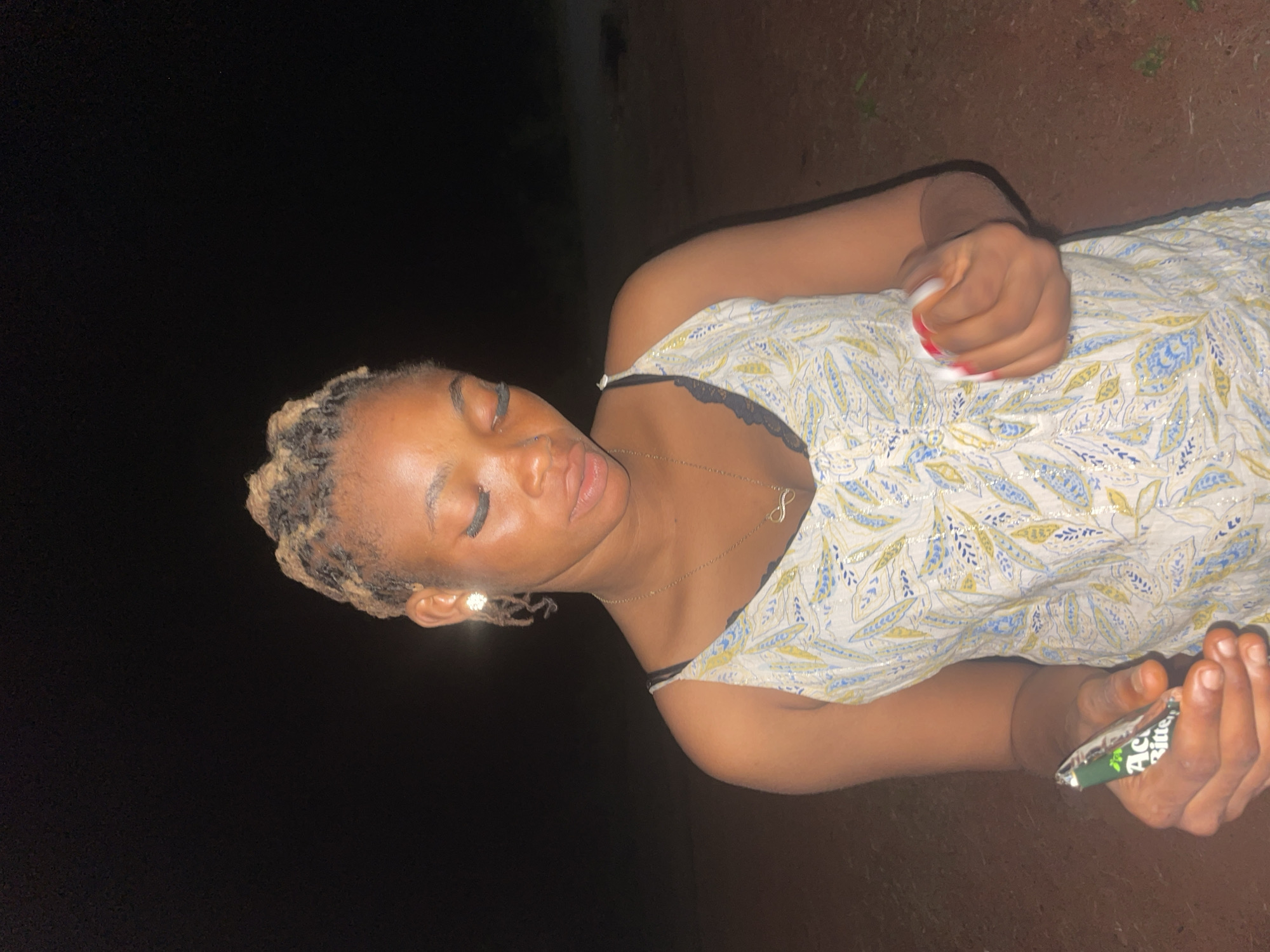 A woman is lying on the ground in a poorly lit area near Ogbe, Nigeria. She has light brown skin, blond dreadlocks, and is wearing a light-colored dress with thin straps, revealing a black bra strap. She is also wearing a gold necklace with an infinity symbol pendant. In her right hand, she holds a white object with red markings, and in her left hand, she holds a green and silver wrapper, labeled with the words "Act Bitter". The ground beneath her appears to be dark soil. The background is dark, suggesting it is night.