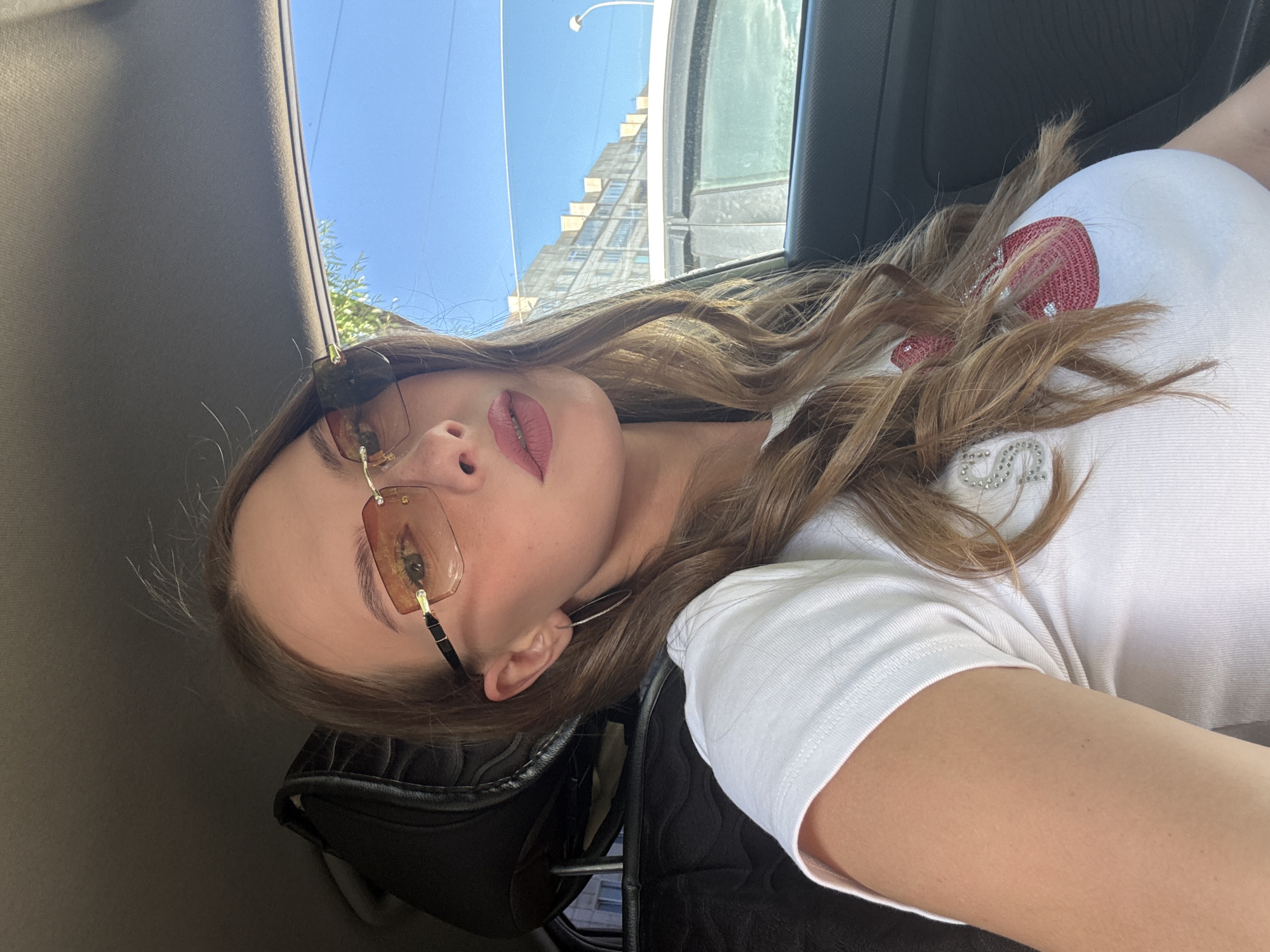 The image captures a young woman indoors, likely in a car, as suggested by the car's interior visible in the background. She is wearing a white t-shirt adorned with red and silver embellishments. The woman is also wearing fashionable rimless sunglasses with brown lenses, which reflect the environment. Her makeup is noticeable, with emphasis on her lips which are a dusty rose color and her eyebrows are well-defined. Her long, wavy, light brown hair is spread across her shoulders.
The scene is illuminated by natural light, indicating it is daytime. The bright sky visible through the window suggests fair weather. In the background, through the car window, a tall building under construction is visible, along with a street lamp. The overall mood conveyed is casual and relaxed, possibly indicating a journey or a leisurely outing. The image is oriented sideways, suggesting it was taken in portrait mode or that the camera was tilted. No visible text is present in the image.