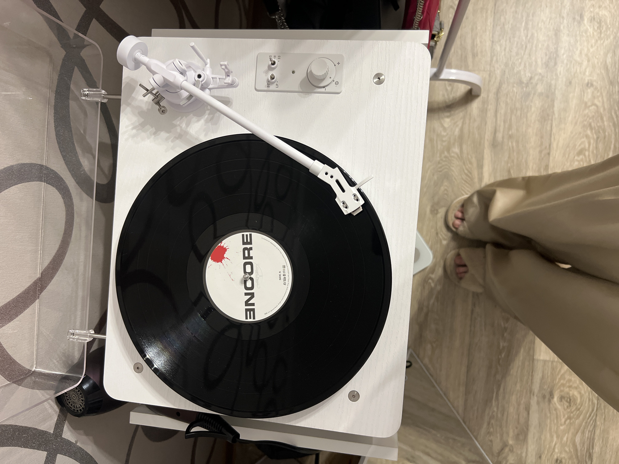 The image shows a close-up, overhead view of a white record player with a black vinyl record. The record has a white label with red splatters and the word "ENCORE" in large, bold letters. A white tonearm is positioned over the record. The record player is on a white stand, which is placed on a wooden floor. In the foreground, the feet of a person wearing light-colored slippers are visible. The background features a wall with a patterned wallpaper. The scene is indoors. There is no information within the image to indicate a specific location in Kazan, Russia.