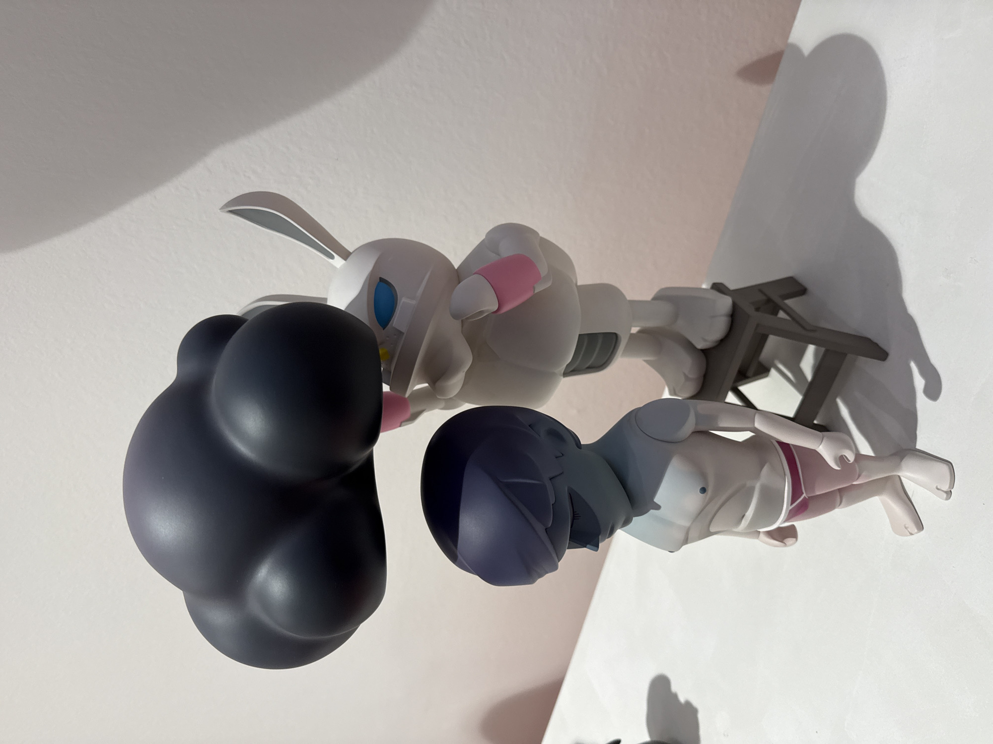 The image shows two toy figures on a white surface against a light pink wall. The figure on the left is a stylized human-like character with a dark blue, sculpted hairstyle and a light gray body wearing pink underwear. Its eyes are closed, and it appears to be looking down. The figure on the right is a white anthropomorphic rabbit-like character with blue eyes and pink accents on its arms. It is standing on a small, gray stool. The rabbit figure is holding a large, dark gray, cloud-shaped object above the head of the human-like figure. The lighting suggests an indoor setting, casting shadows on the white surface and the wall. The overall scene is artificial and staged, typical of an art exhibit or display. No specific location in Moscow, Russia, can be determined from the image.