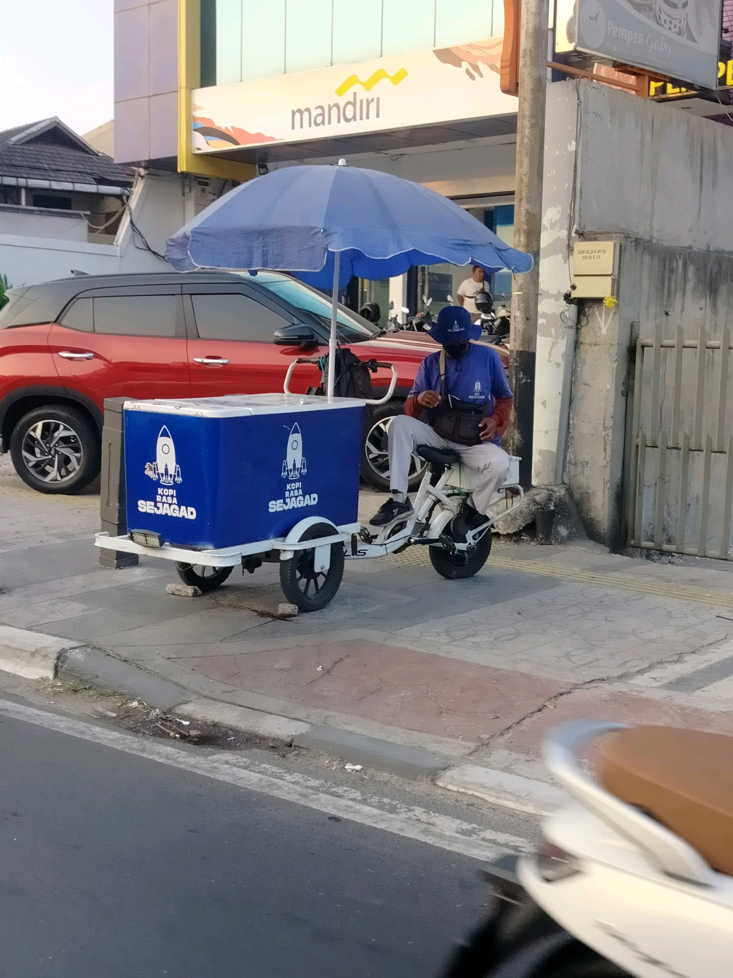In East Jakarta, Indonesia, a vendor sits on a white electric scooter attached to a blue ice chest cart. The cart is adorned with white rocket graphics and the text "KOPI RASA SEJAGAD". The vendor, wearing a blue t-shirt, light-colored pants, a blue hat, and a black face mask, is looking down at a black bag. A large blue umbrella shades the cart and the vendor. Behind the cart is a red SUV, and further back, a building with a yellow and blue sign that reads "mandiri". The scene is on a paved sidewalk next to a street.