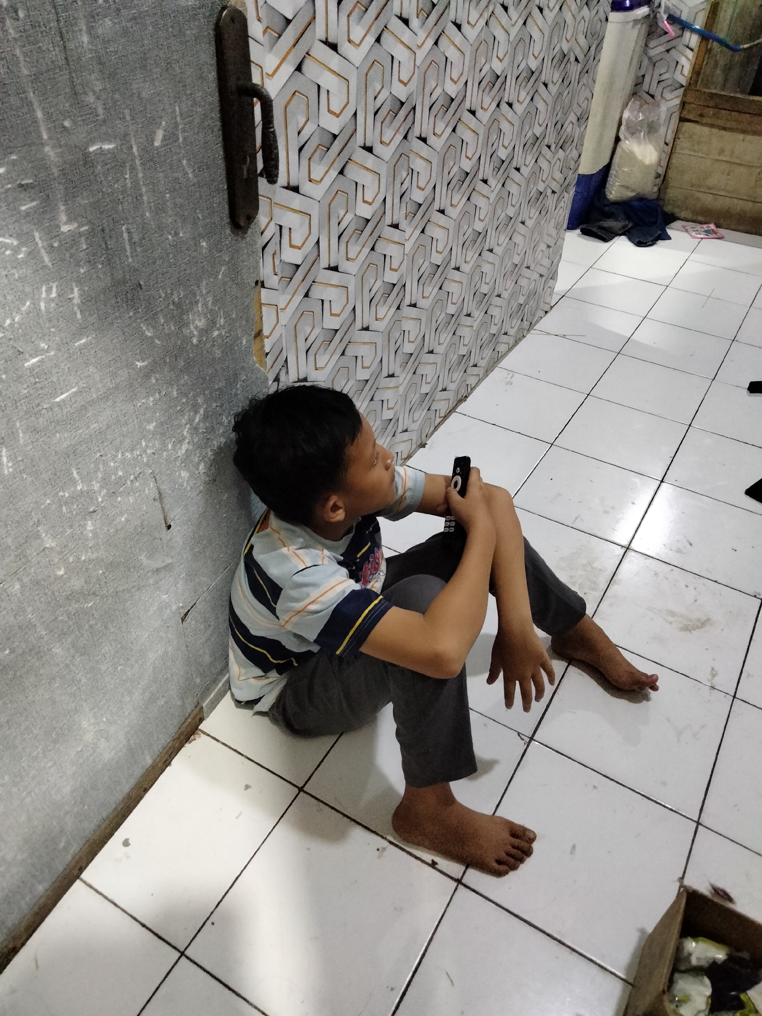 A young boy is sitting on a tiled floor, leaning against a gray wall with peeling wallpaper and a metal door handle. He is wearing a striped t-shirt and dark pants, and holding a black remote control. To his right, the wall is decorated with a geometric wallpaper pattern. In the background, there are some stored items, including a large blue cylindrical object, a plastic bag, and some dark fabric. The floor tiles are white with dark grout lines, and some appear to be stained. The scene appears to be indoors, possibly in a residential area in Cariu, Indonesia.