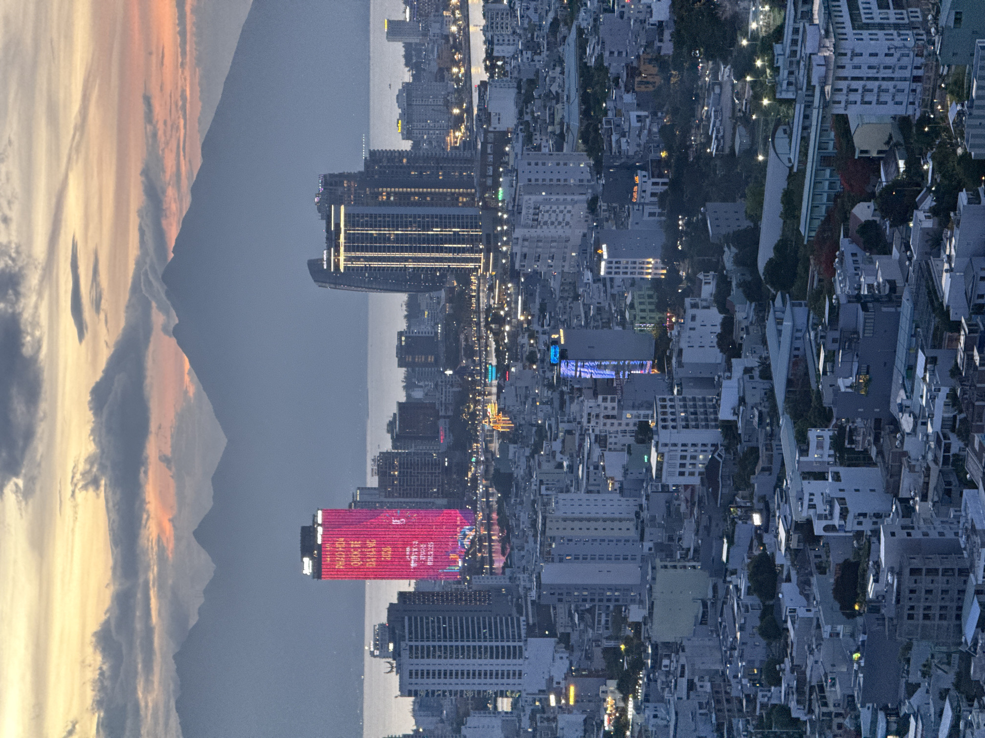 This image presents an expansive urban landscape at dusk or night, showcasing a city nestled alongside a body of water with mountains in the background. The scene is viewed from a high vantage point, looking across the illuminated cityscape.
The upper portion of the image reveals a dramatic sky filled with scattered clouds, displaying a gradient of warm golden and orange hues on one side transitioning to darker, cooler tones. Silhouetted against this sky are prominent mountain ranges, indicating the city's geographical setting.
The city itself is a dense collection of buildings of varying heights and architectural styles. Many buildings are brightly lit, with windows glowing and streetlights forming intricate patterns across the urban fabric. A significant body of water, likely a river or the sea, runs through or alongside the city, reflecting some of the artificial lights.
A striking feature is a tall, modern skyscraper illuminated with a vibrant red light. White vertical text is displayed on its facade, clearly visible as "DANANG SUMMER FESTIVAL," with "PREMIUM" also partially legible above it. Another notable building nearby features prominent vertical golden light strips. The surrounding urban areas are a mosaic of rooftops, green spaces, and more residential-style buildings.
The overall impression is a bustling metropolis alive with light after sunset, set against a backdrop of natural grandeur. The scene depicts Đà Nẵng, Vietnam, at night, highlighting its urban development and natural beauty. No individual people or specific activities are discernible, but the illuminated city implies widespread human presence and ongoing urban life.