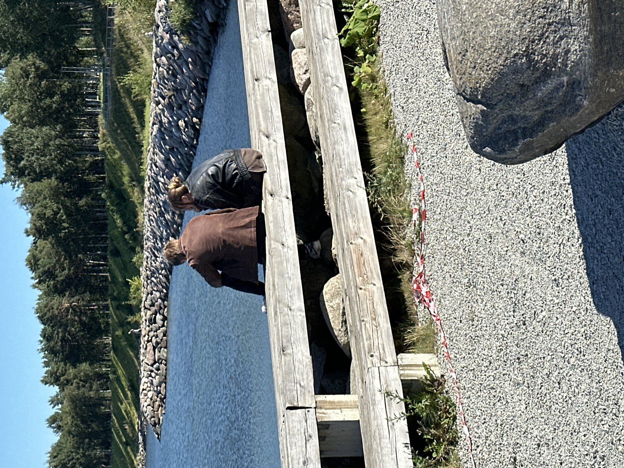 Two individuals, a male and a female, sit on a wooden railing overlooking a body of water. The male, on the left, wears a brown jacket and dark pants. The female, to his right, is dressed in a black leather jacket and brown trousers. They are positioned with their backs to the camera.
The railing, constructed from weathered wooden planks, spans across a rocky embankment that borders a tranquil blue body of water. Behind the water, a dense line of green trees forms a backdrop against a clear blue sky. The ground in the foreground is covered in small gray gravel. Red and white caution tape is visible on the gravel area, near the railing, suggesting a restricted or work in progress zone.
The scene appears to be a public area, possibly a park or promenade, situated along the coast of Tallinn, Estonia. The presence of the rocky embankment and the expansive water suggests a coastal location. The clear skies and sunny weather indicate a pleasant day. No specific landmarks or identifiable features of Tallinn are clearly visible to pinpoint the exact location within the city, but the overall environment is consistent with the city's coastal geography.
