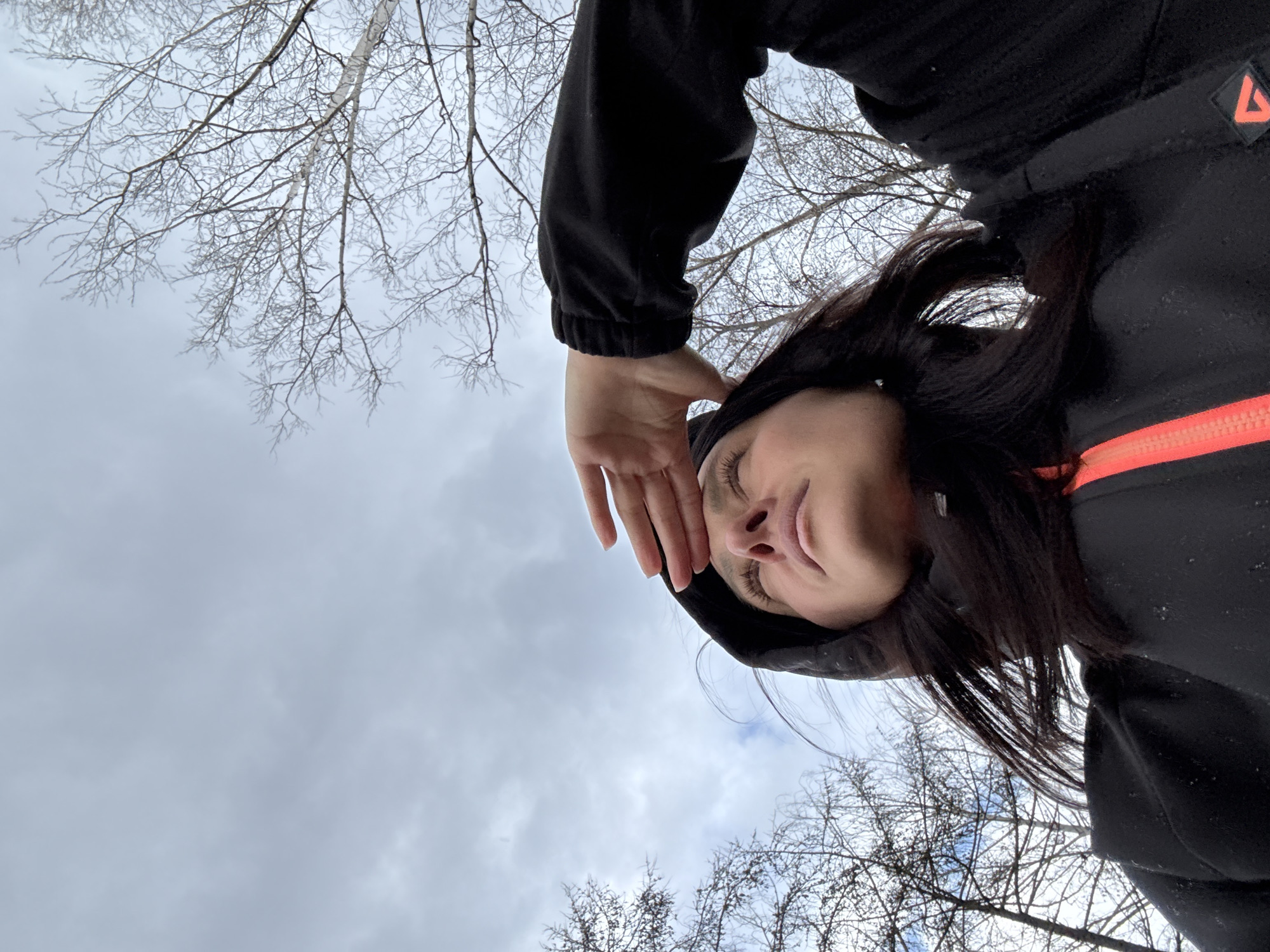 The image presents an upward-angled perspective of a person outdoors against a cloudy sky and bare tree branches, consistent with a cold climate like Chaikovsky, Russia.
The main subject is a person with long dark hair, seen from below. They are wearing a black hooded jacket, which stands out with a bright orange vertical zipper and a small, orange triangular logo on the upper right sleeve. The person's right hand is raised to their forehead, partially obscuring their face, which shows a subtle, perhaps contemplative, expression. Their eyes are visible, looking slightly upwards.
The background is dominated by an overcast sky, filled with varying shades of light grey clouds. Numerous bare, intricate branches of deciduous trees are scattered throughout the frame, suggesting winter or early spring. Some branches appear to have a delicate dusting, possibly frost or snow, highlighting the cold weather.
The activity appears to be an outdoor moment, possibly a self-portrait or a low-angle photograph taken by another person. The scene conveys a quiet, cold, and possibly serene atmosphere. No specific time of day is discernible due to the overcast conditions, but it is clearly daytime. No visible text is present other than the indistinct triangular logo on the jacket.