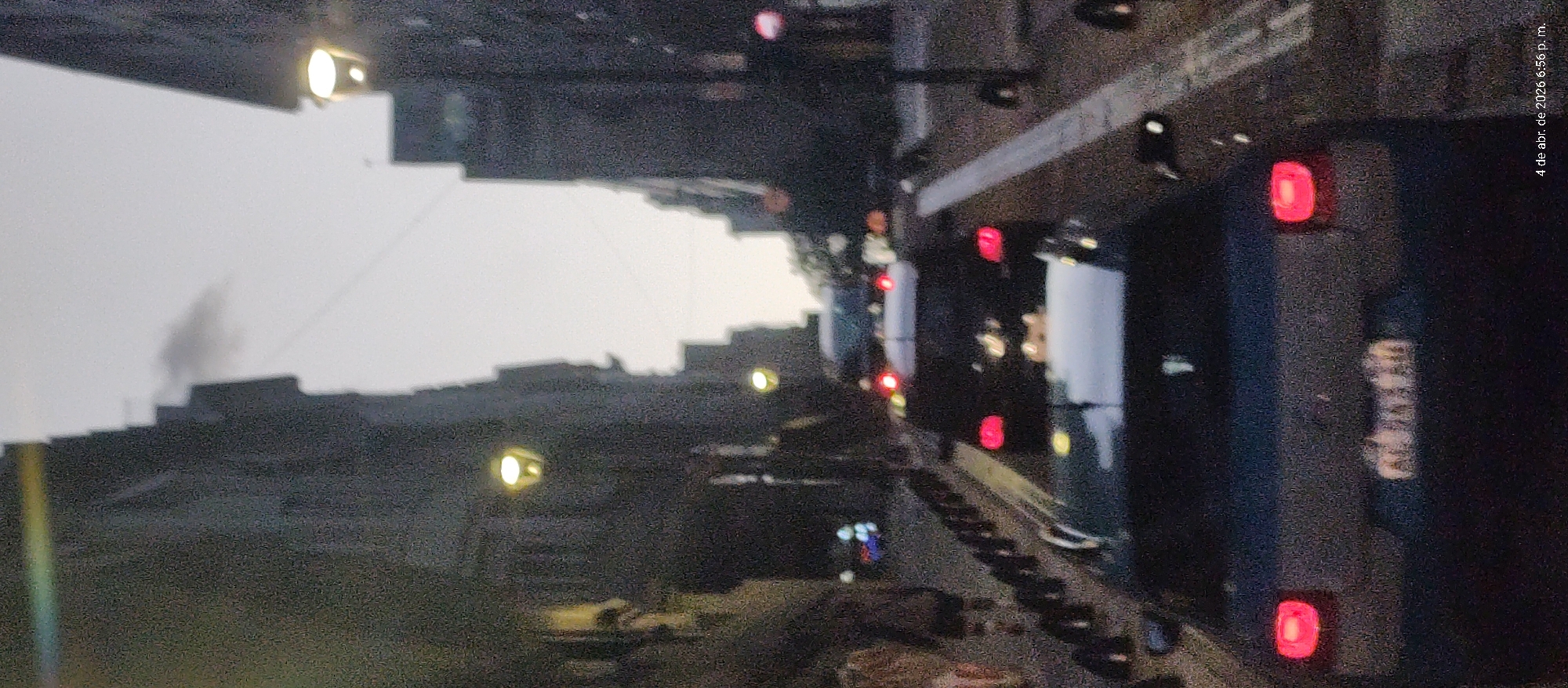 Here is a description of the image:
The image shows a nighttime urban scene. Buildings line the left side of the frame, punctuated by streetlights. On the right side, traffic is visible with car taillights and license plates. The photograph was taken on April 4, 2026, at 6:56 p.m. and is possibly located in Buenos Aires, Argentina.