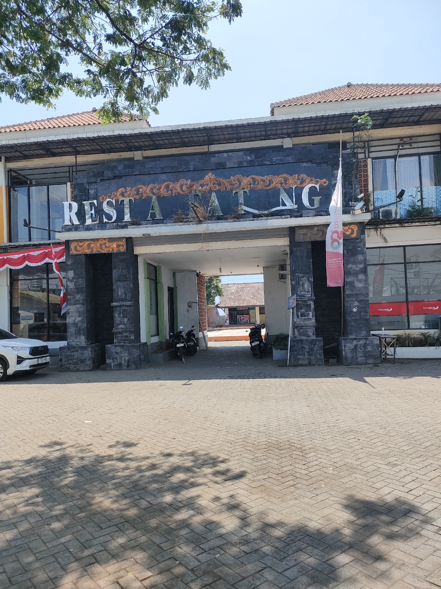 A wide shot captures the facade of a building in Curahdringu, Indonesia, likely a restaurant given the prominent "REST A NG" signage in white lettering against a dark, textured wall. The building features a covered entrance archway with dark, stone-like pillars. On the left, a white SUV is parked in front of a window display draped with red and white fabric. Motorcycles are visible parked under the archway and to the right of it. A red and white flag with text hangs from a pole on the right side of the entrance. The ground in front of the building is paved with interlocking bricks, and shadows from overhead foliage are cast across it. The sky is clear and blue, suggesting a sunny day. The architecture suggests a mix of modern and traditional elements. The overall scene depicts an urban commercial area.