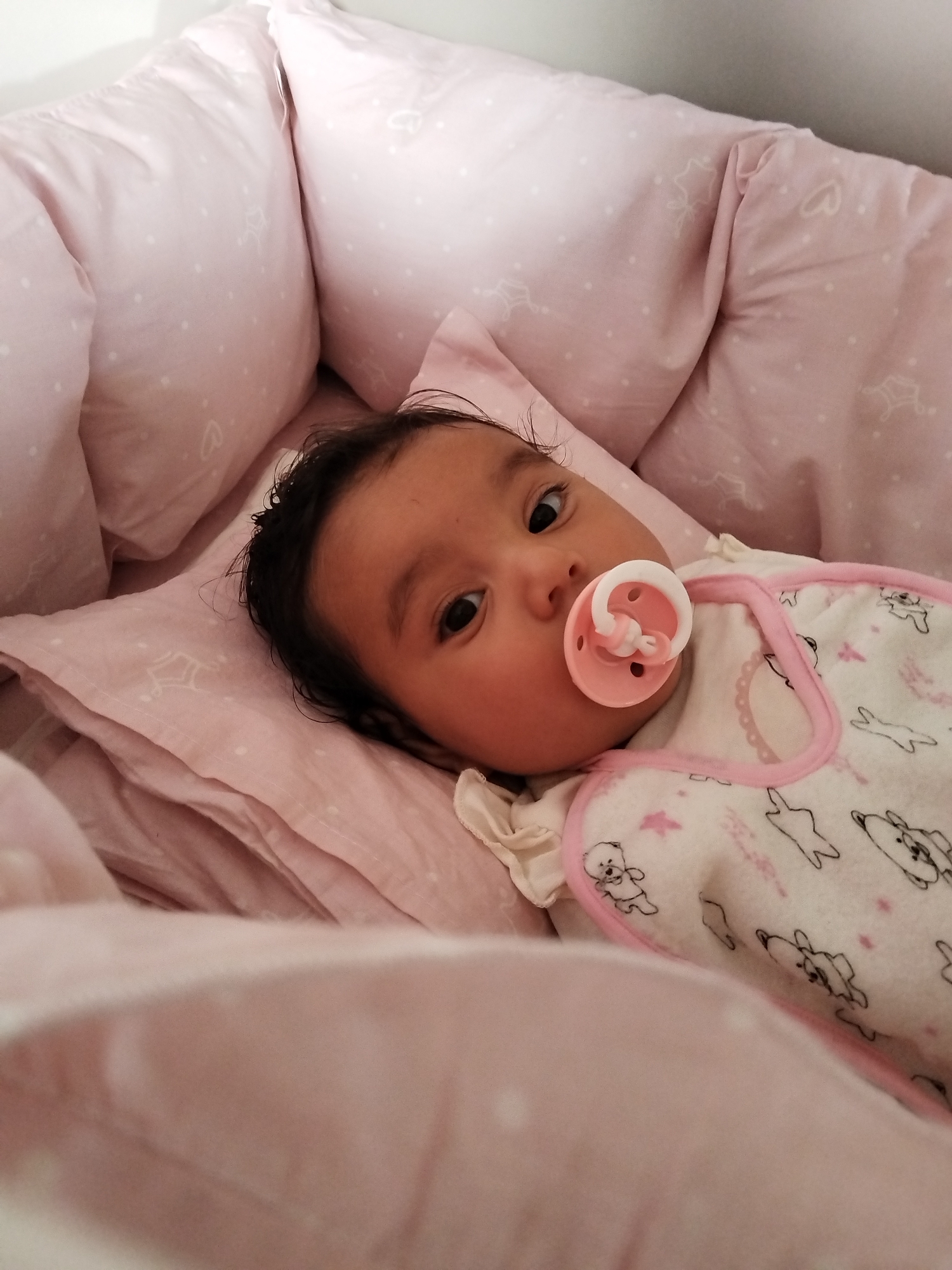 A baby is lying in a bed surrounded by pink bedding with printed stars and shapes. The baby has dark hair and is using a pink pacifier. The baby is wearing a white sleeper with pink trim and a cartoon pattern. The location is unknown.