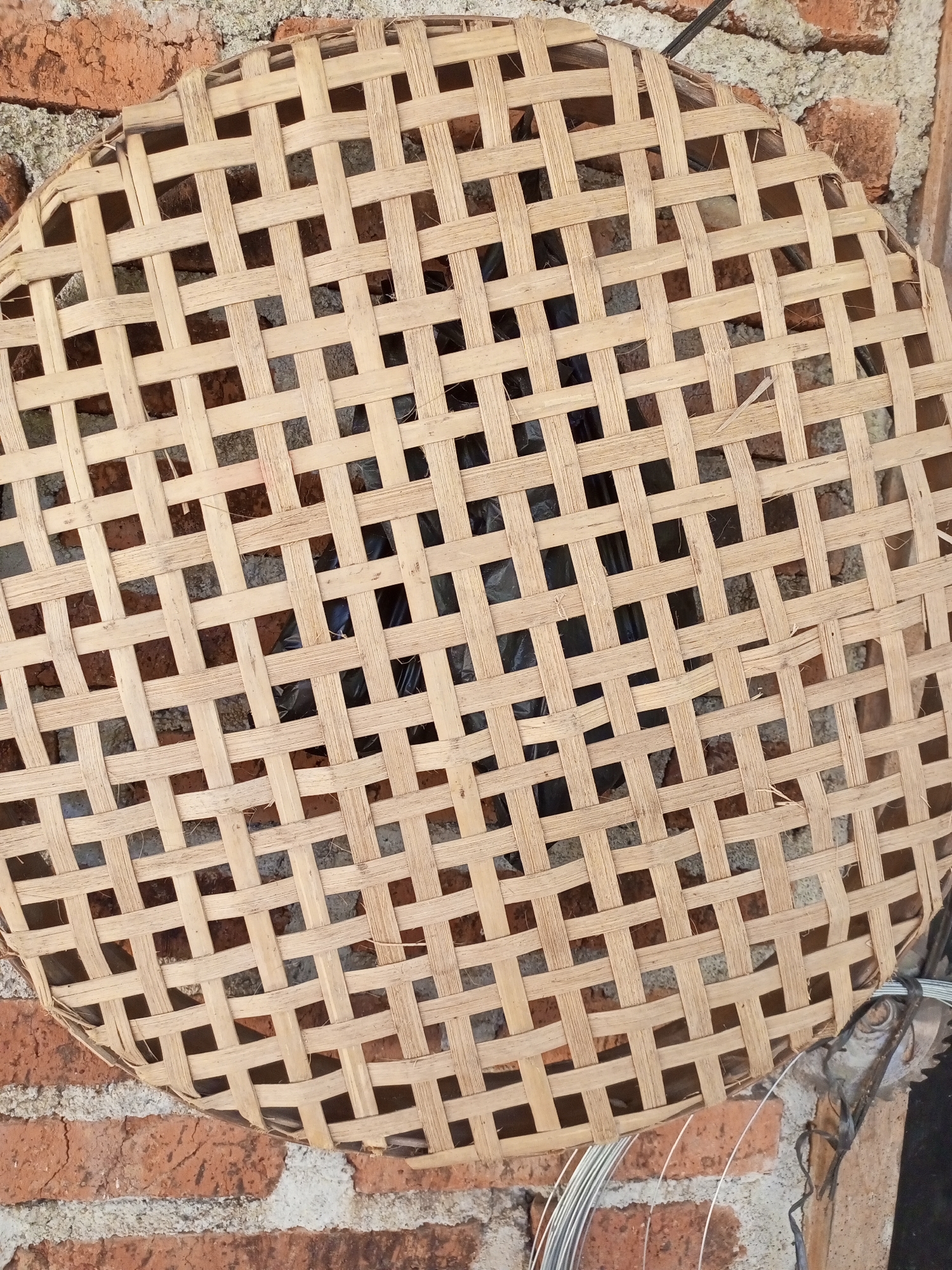 A close-up, overhead shot shows a circular woven bamboo tray, or "tampah," resting against a rough brick wall. The bamboo strips are light brown and interwoven in a grid pattern, creating square openings through which dark objects and the textured brick wall are visible. The tray is slightly warped, with some edges appearing frayed. The brick wall behind it is a mix of reddish-brown bricks and lighter colored mortar or concrete. To the right of the tray, a bundle of thin metal wires is partially visible, along with some dark string or cord. The scene appears to be an outdoor setting, possibly a yard or storage area, in Jatipurno, Indonesia, given the context. There are no people or animals in the image.