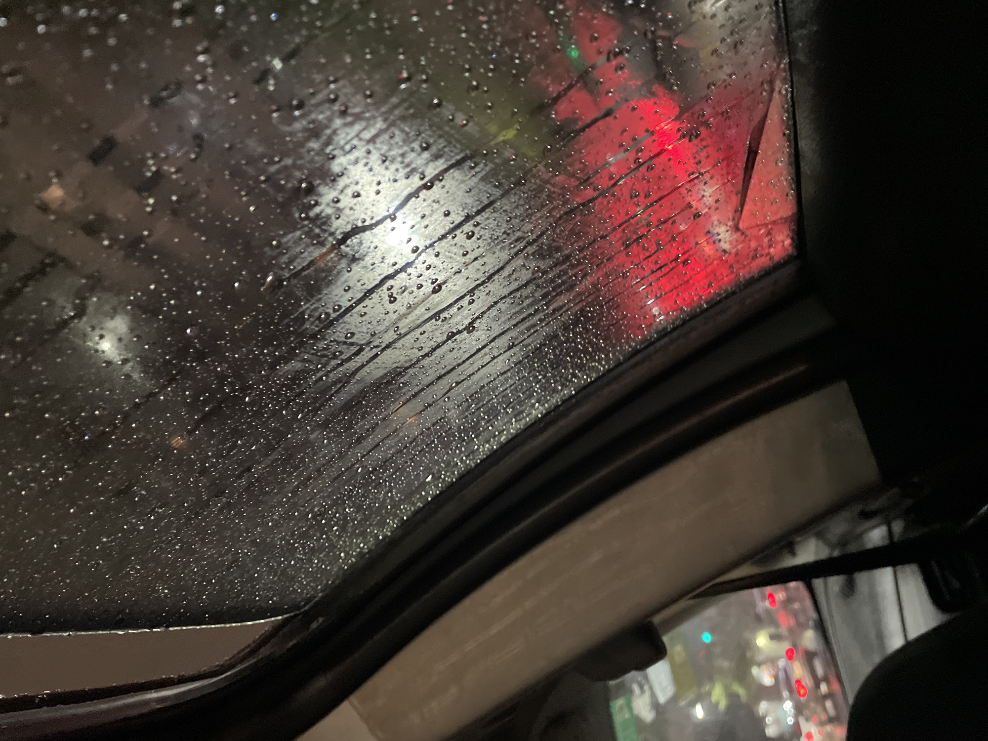 The image is taken from inside a car looking through the sunroof during a rainstorm. The glass is covered in water droplets. Outside, blurred lights suggest a city street at night. Red brake lights and other ambient light sources are visible. The interior of the car, including the frame of the sunroof and part of a seat, are also visible. It is not possible to identify the specific location in Buenos Aires from the image. There are no visible people, actions, or interactions directly discernible.