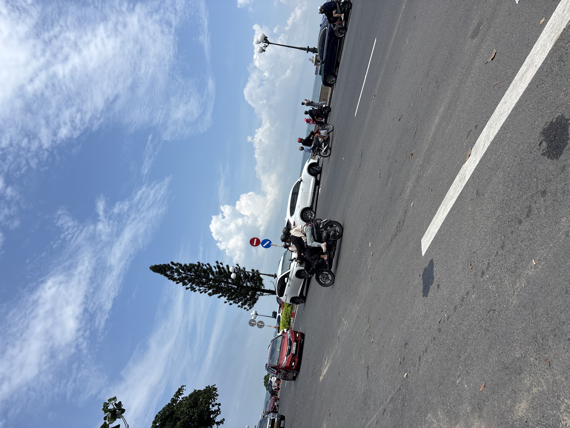 A wide-angle shot captures a coastal road in Ho Chi Minh City, Vietnam, on a partly cloudy day. The road is asphalt with white lane markings. In the foreground, a black motorcycle with two passengers, wearing helmets, is traveling to the right. To the left of the motorcycle, a red car is also moving to the right, followed by other vehicles. Further down the road, a white SUV and a dark sedan are visible. Along the right side of the road, a lamppost with multiple lights stands near the sea. The background features a body of water, likely the ocean, and a lush green tree with a tall, slender evergreen tree in the middle ground. The sky is a bright blue with scattered white clouds. The overall scene depicts a typical day on a busy coastal road with traffic and pedestrians.
