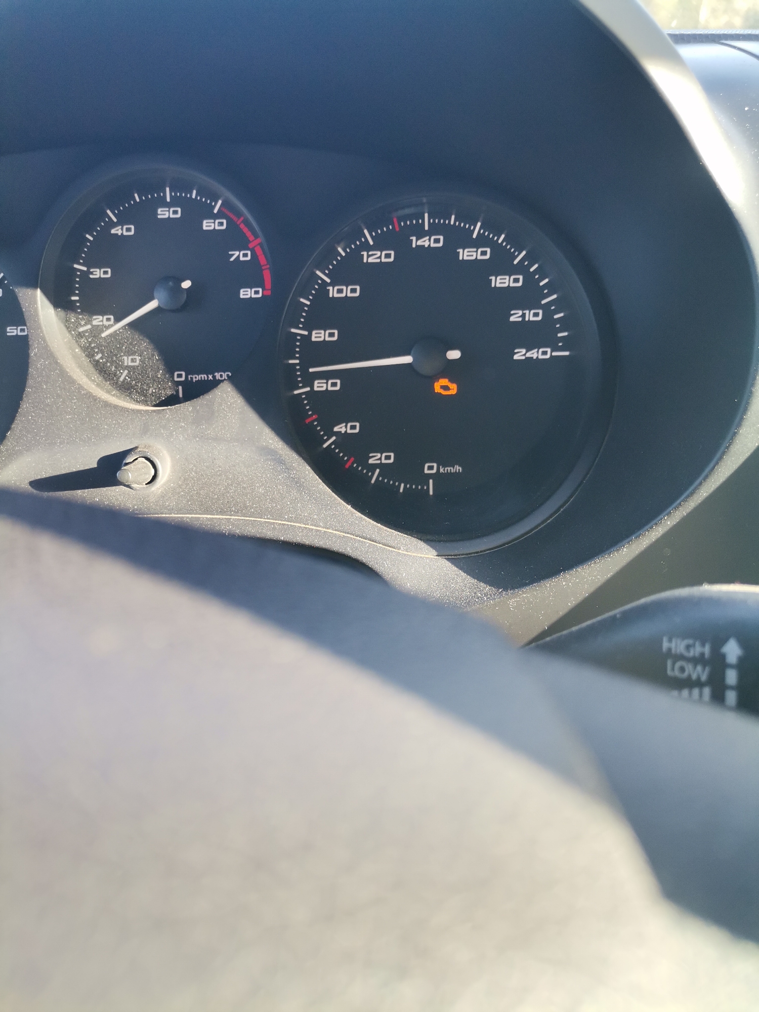 The image displays a close-up view of a car's dashboard, specifically the instrument cluster, from the driver's perspective, likely captured during daylight hours in Batovo, Bulgaria. The interior surfaces of the dashboard and instrument panel show a noticeable layer of dust.
The left gauge is a tachometer, indicating the engine is running at approximately 2000-2500 RPM (reading between '20' and '30' on the 'rpm x 100' scale). The right gauge is a speedometer, showing the vehicle is currently traveling at approximately 80 km/h. The speedometer's maximum reading is 240 km/h.
A prominent detail is the illuminated orange "Check Engine" warning light visible beneath the speedometer needle, indicating a potential issue. Part of a control stalk is visible on the right, featuring text "HIGH" and "LOW" and an upward arrow, likely controlling headlights or wipers. A small adjustment knob or button is located on the lower left of the instrument cluster. The bright sunlight creates reflections and shadows across the dashboard.
Visible text includes "rpm x 100", numerical values "10" through "80" on the tachometer, and "km/h" with numerical values "20" through "240-" on the speedometer. The control stalk also clearly displays "HIGH" and "LOW".