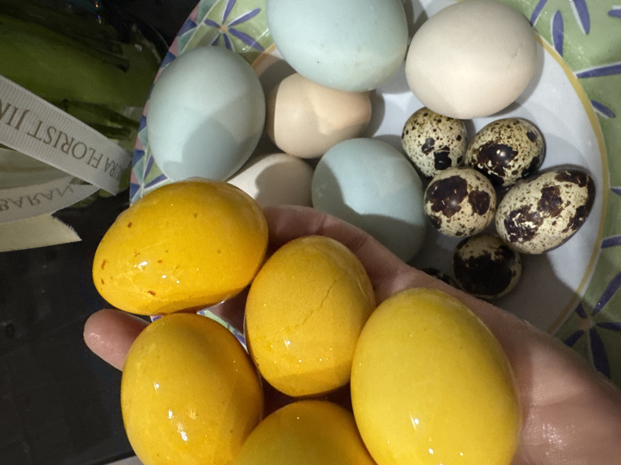 A hand holds several yellow eggs, likely dyed. A plate in the background contains a variety of eggs, including light blue, beige, white, and quail eggs. A bottle with florist ribbon is visible to the left. The location is unknown.
