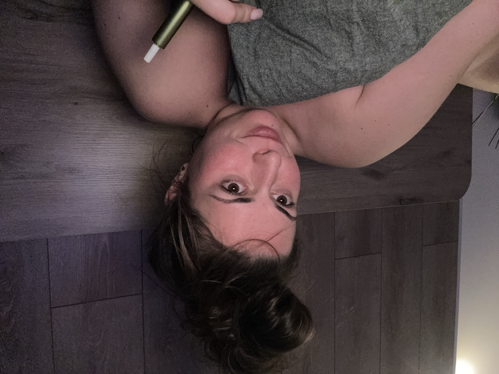A woman with her hair in a messy bun is lying in bed, covered by a gray blanket. She is holding a metallic green electronic cigarette in her right hand, with her index finger extended towards the tip. The woman has fair skin, brown eyes, and rosy cheeks. She is looking towards the camera with a slight smile. The background consists of a dark wood-paneled wall and a headboard. The lighting is dim, suggesting a nighttime setting. The woman is wearing a strapless garment.