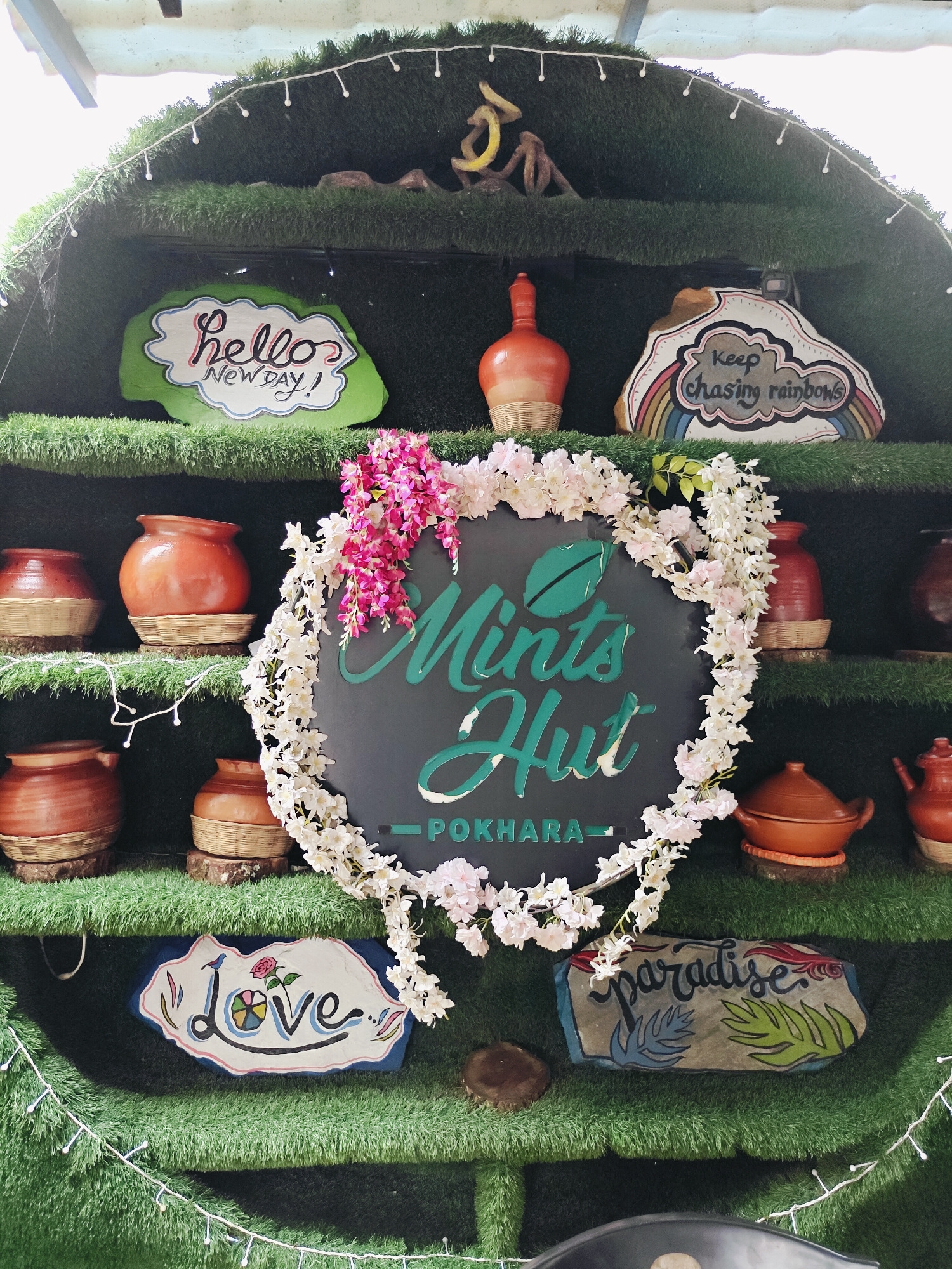 The image depicts a rustic display likely located at Mints Hut in Pokhara, Nepal. The display is arranged on shelves covered in artificial green grass, resembling a tiered shelf or a decorative wall.
On the top shelf, a green, cloud-shaped sign reads "Hello NEW DAY!" in stylized lettering. To its right, a white, irregular-shaped sign features a rainbow and the text "Keep chasing rainbows." A small, orange, terracotta pot sits between these signs.
The middle section of the display is dominated by a large, round, dark green sign with the words "Mints Hut POKHARA" in a lighter green, cursive font. This sign is adorned with a floral garland of pink and white artificial flowers. To the left of this central sign are four terracotta pots, each with a woven base, arranged in two rows. To the right, a similar terracotta pot is visible, and further to the right, a small terracotta teapot.
The bottom shelf features two more decorative signs. On the left, a white sign in the shape of a lip has the word "Love" painted on it, with a small bird and a rose design. On the right, a rectangular stone has the word "paradise" written on it, accompanied by stylized leaf motifs. Additional terracotta pots are visible on the shelves, and white fairy lights are strung along some of the shelves, adding a festive touch. The overall impression is that of a charming and decorative entrance or display area for a café or eatery.