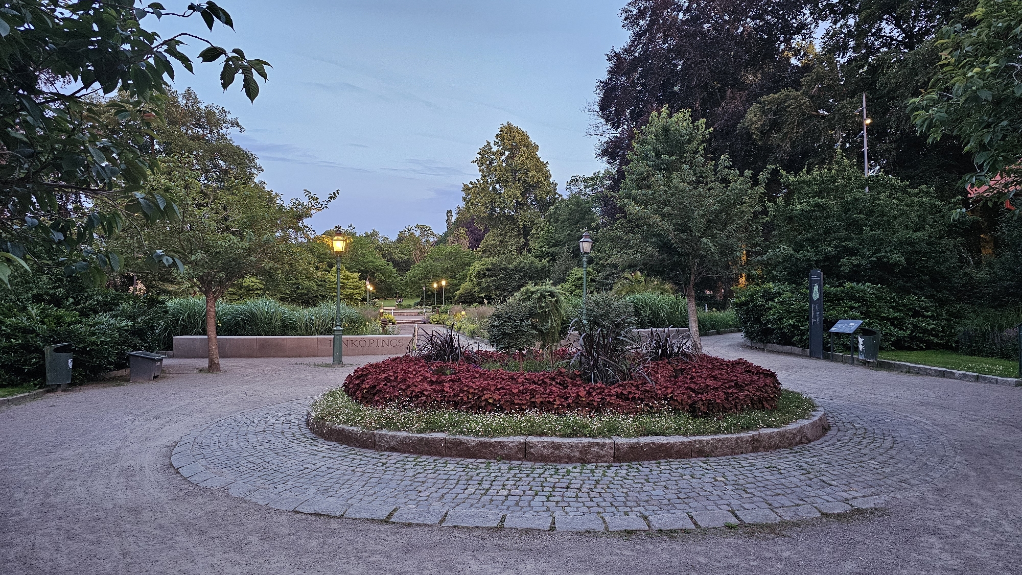 The image shows a park in Linköping, Sweden, with a circular flower bed in the foreground filled with red foliage and white flowers. The flower bed is surrounded by a cobblestone path. In the background, there are trees, lampposts, and a stone wall with text that reads "LINKÖPINGS". The sky is overcast. There are no people or animals visible in the image. The overall scene is peaceful and serene.
