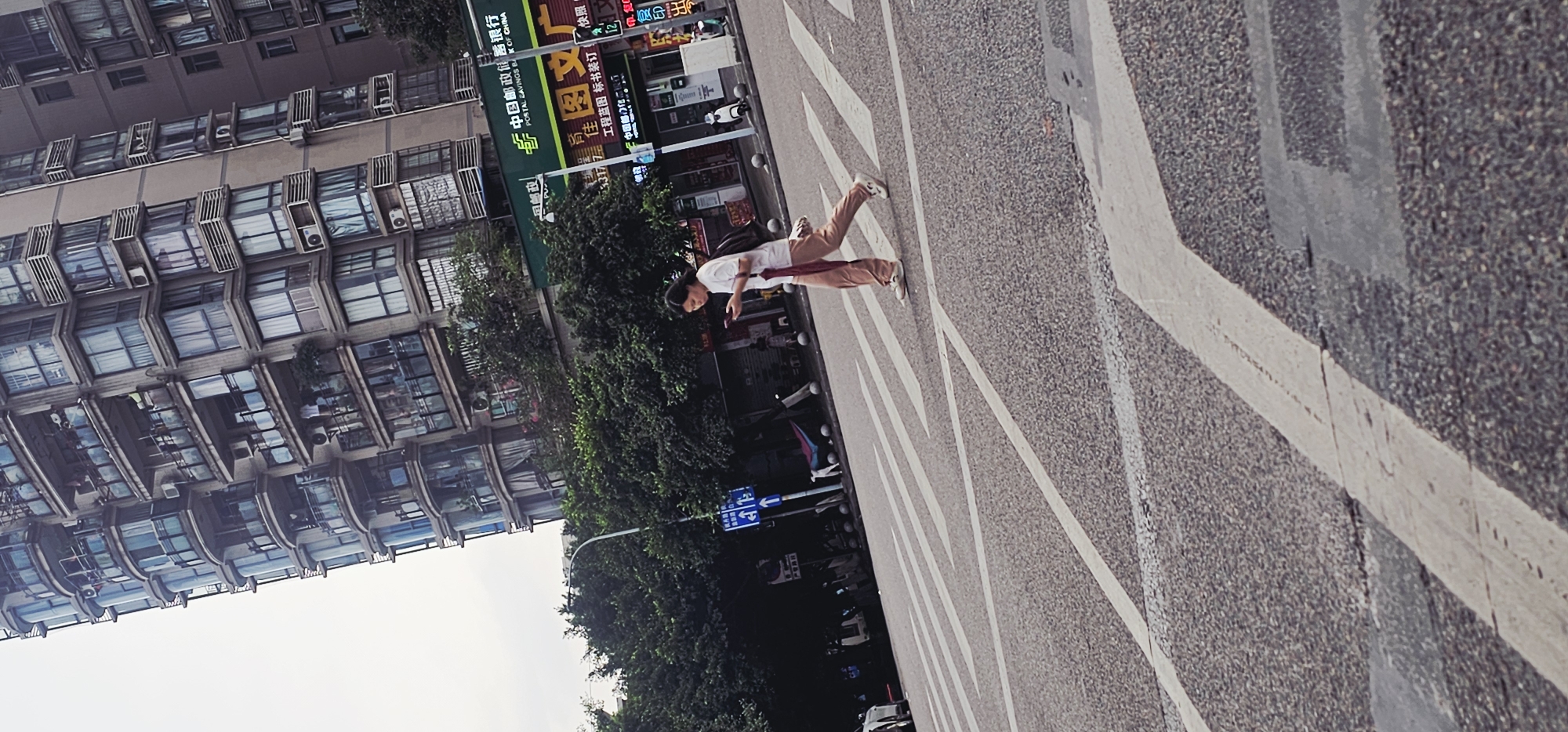 The image shows a woman walking across a crosswalk in Jiulongpo District, China. She is wearing a white top and light brown pants and appears to be looking at her phone. Behind her is a building with many windows and air conditioning units. There are trees and a street sign visible. A "China Postal Savings Bank" branch and other stores are in the background.