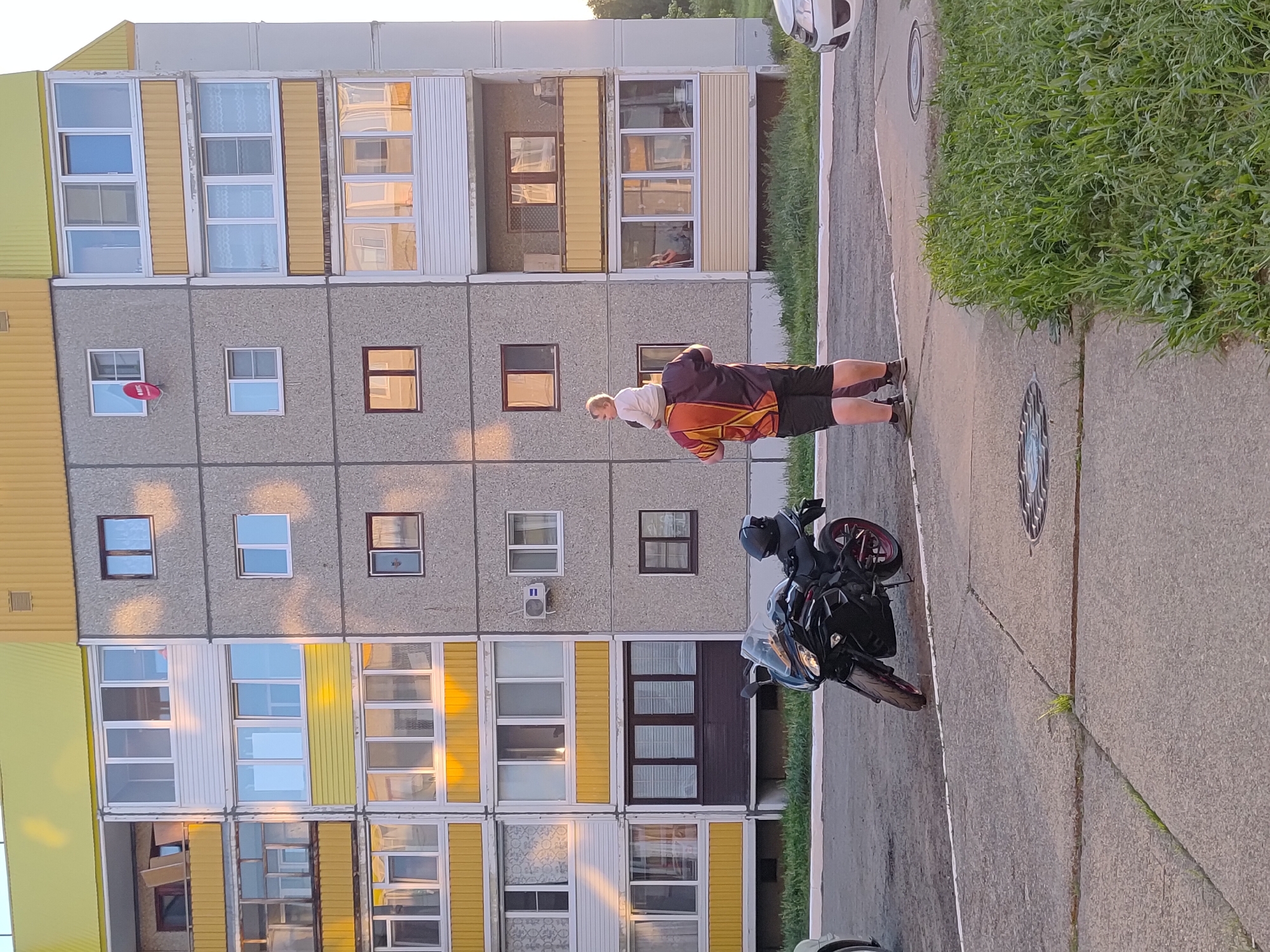 A photograph taken in the city of Марковский, Russia, shows a man carrying a child on his back near a parked black motorcycle, positioned in front of a multi-story building. The building has a repeating pattern of grey concrete panels punctuated by windows, with yellow and white accents framing the windows. The sidewalk is concrete and adjacent to a grassy area.