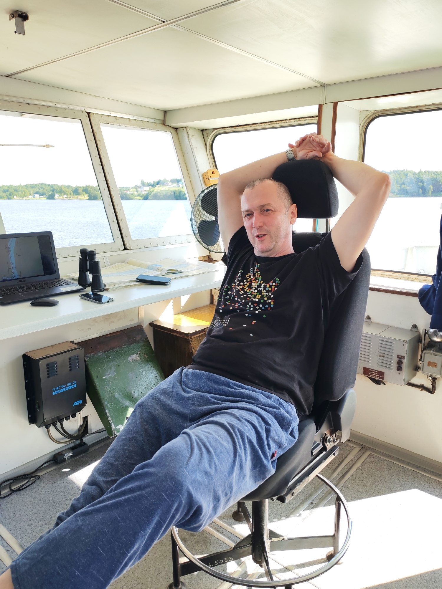 A bald, middle-aged man is seated in a captain's chair inside a ship's bridge, with his arms resting behind his head. He is wearing a black t-shirt with a colorful, abstract pattern and blue sweatpants. The bridge has large windows that offer a view of a body of water with trees and houses on the shore. On the desk in front of him are a laptop, binoculars, a map, and two cell phones. A black fan is visible to his right, and a power supply unit is mounted on the wall to his left. The scene is brightly lit by natural sunlight.