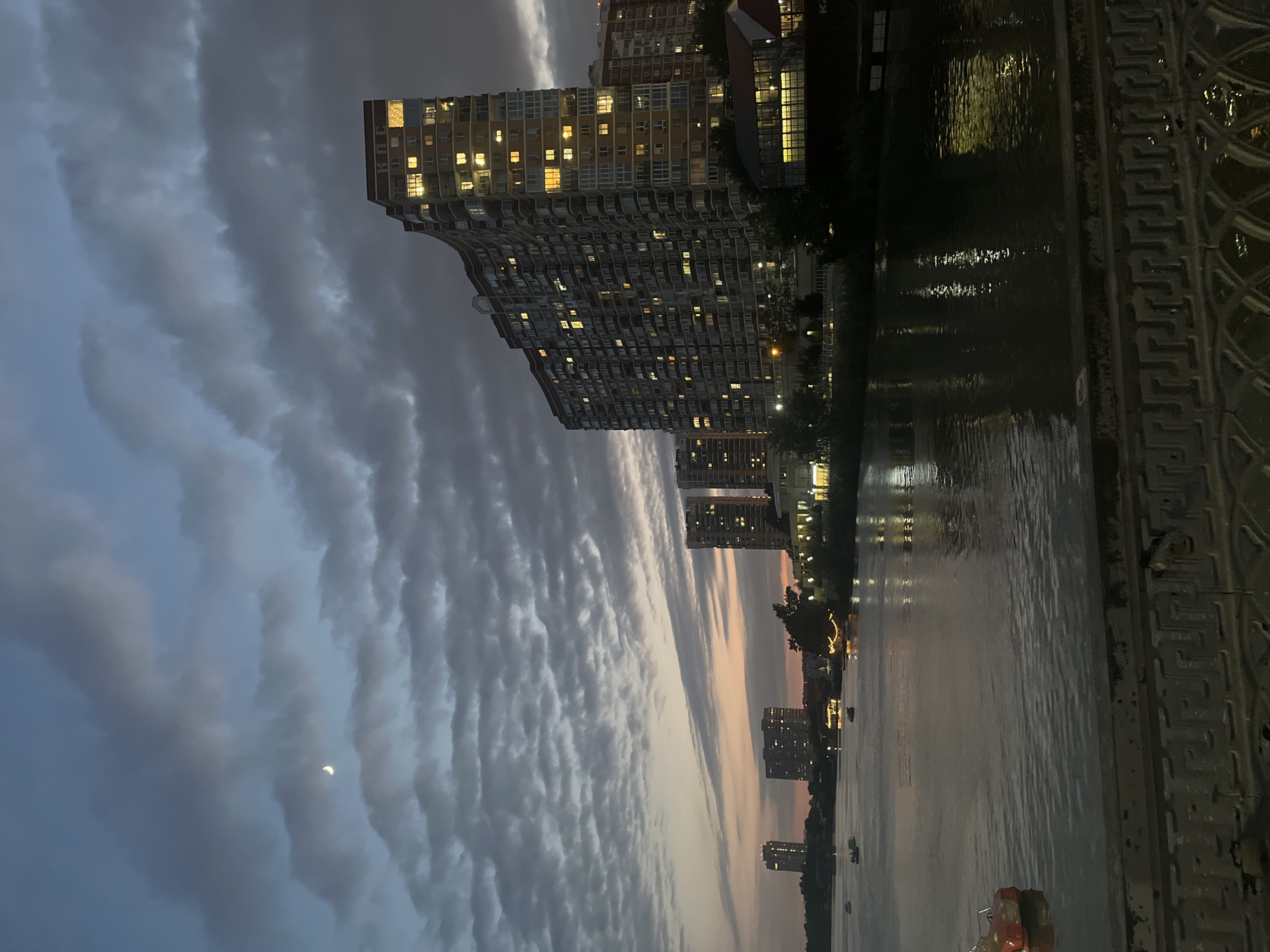 The image depicts a twilight scene along a river in Krasnodar, Russia, likely near the Kuban River embankment. Several multi-story residential buildings are visible on the opposite bank, with lights illuminated in many of their windows, suggesting it is evening. One prominent building is curved and appears to be a modern apartment complex. The sky is cloudy with hints of a sunset, and a sliver of the moon is visible. The river's surface reflects the lights from the buildings and the sky. Small boats are scattered on the water, some appearing to be pedal boats. In the foreground, a decorative metal railing suggests the image was taken from a bridge or elevated walkway. The overall atmosphere is calm and urban.
