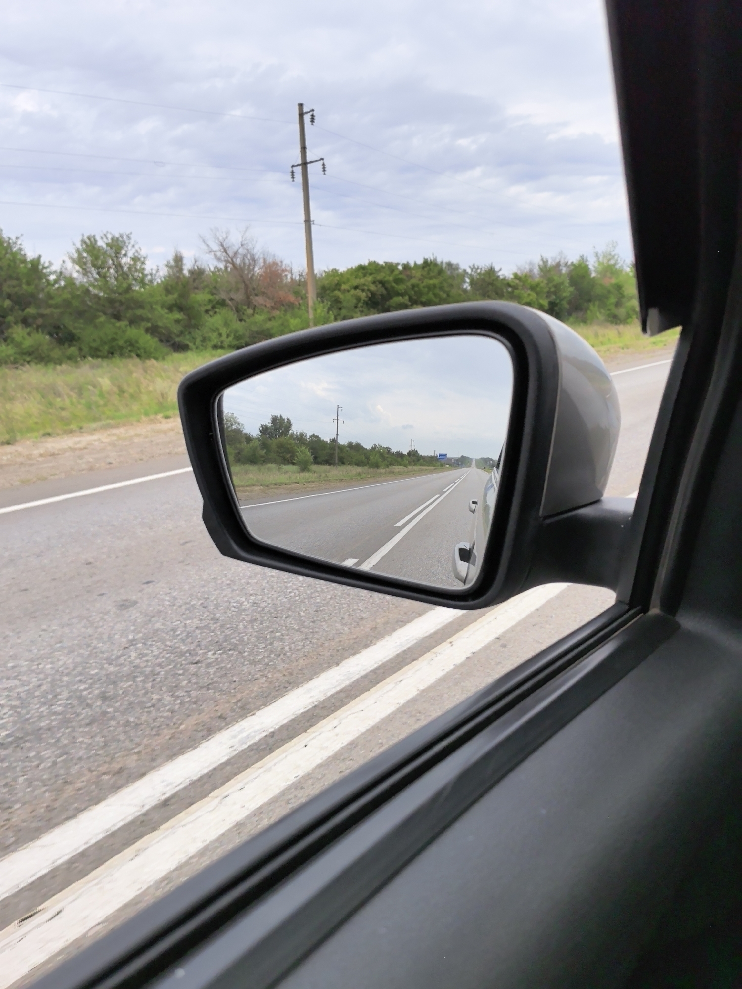 The image shows a view from inside a car, looking at the side-view mirror. The mirror reflects a road with trees and a utility pole in the background. The car is in motion, as indicated by the blur of the road markings and the surrounding scenery. The sky is overcast with clouds. The image is taken from the perspective of a passenger, looking out the right side of the car.